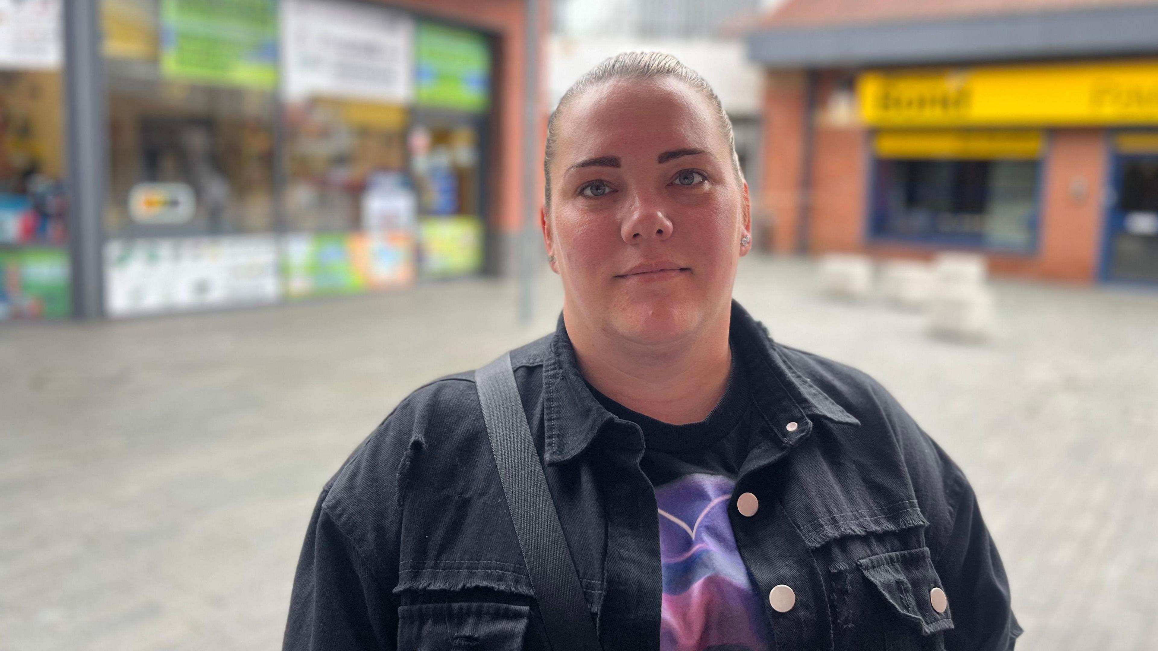 Carina Bradbury has dark hair tied back in a pony tail and is wearing a dark navy denim jacket over a purple and black top. A black bag strap is over her right shoulder. She is standing in a cobbled square with shops behind her.