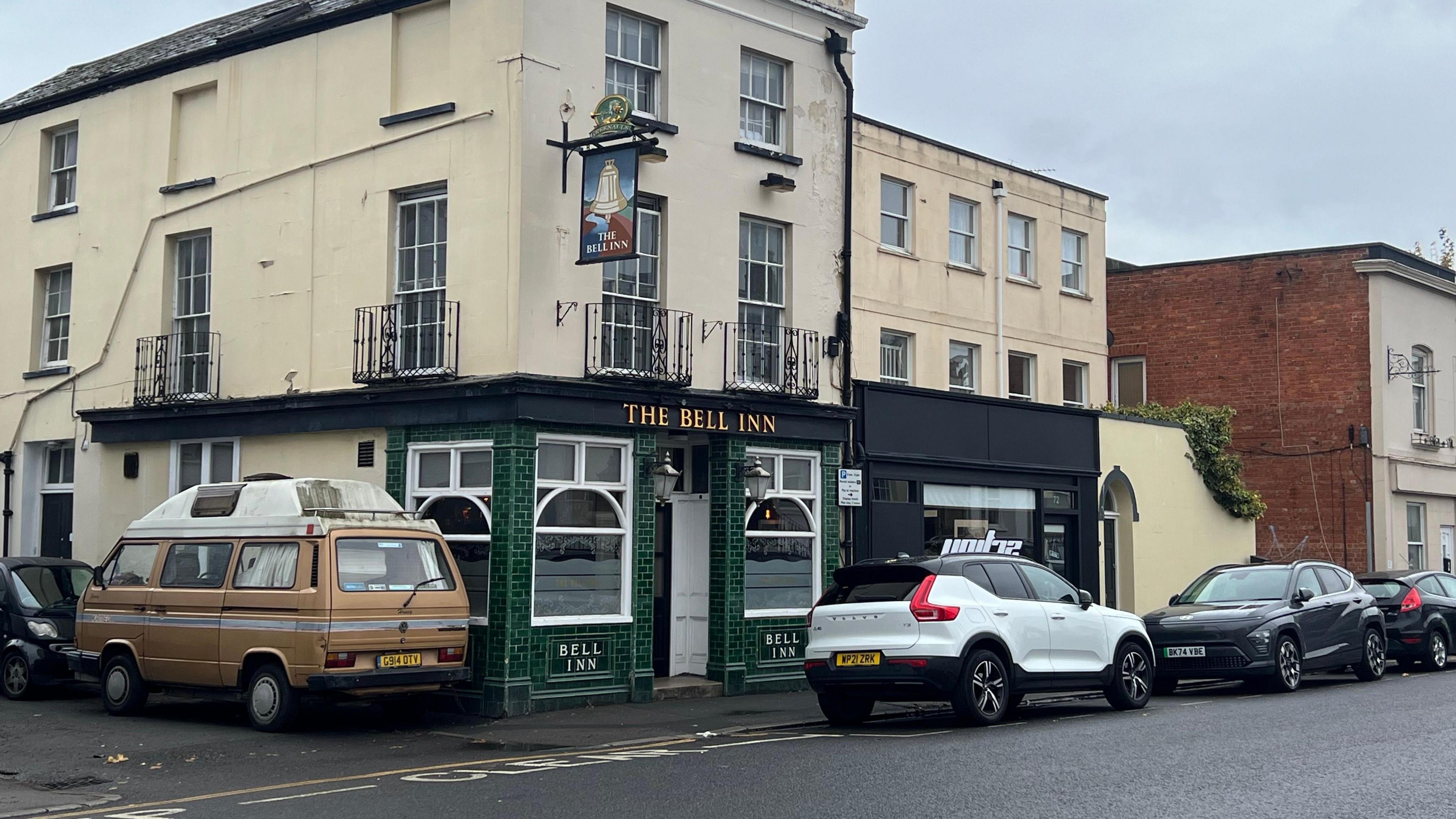 Exterior shot of The Bell Inn taken on a cloudy day. The pub is three storeys and the front entrance is dark green. There are several cars parked outside it.