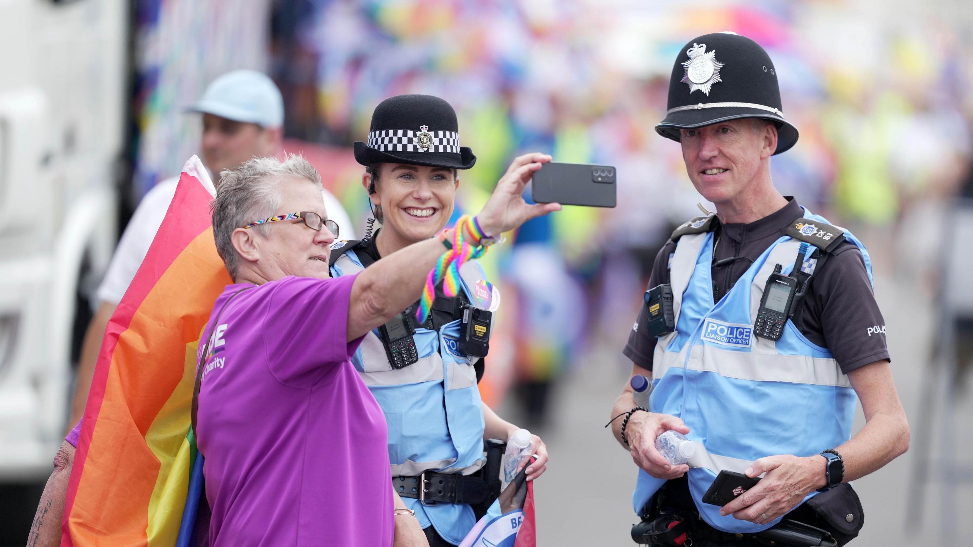 Police liaison officers in blue vests taking a selfie with a woman during the parade. The elderly woman has a purple shirt and is carrying a rainbow flag