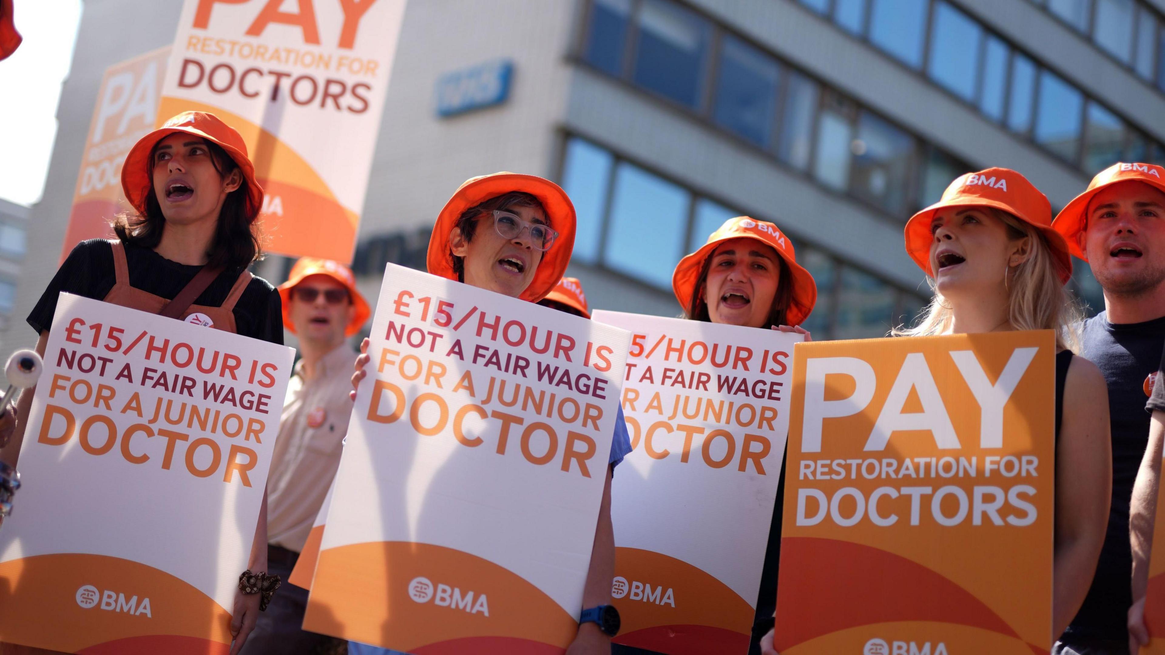 A group of junior (now called resident) doctors holding placards outside a hospital in London last year. They are holding signs saying "£15 an hour is not a fair wage for a junior doctor" and wearing orange hats with the BMA logo on them. They look like they are singing or chanting.