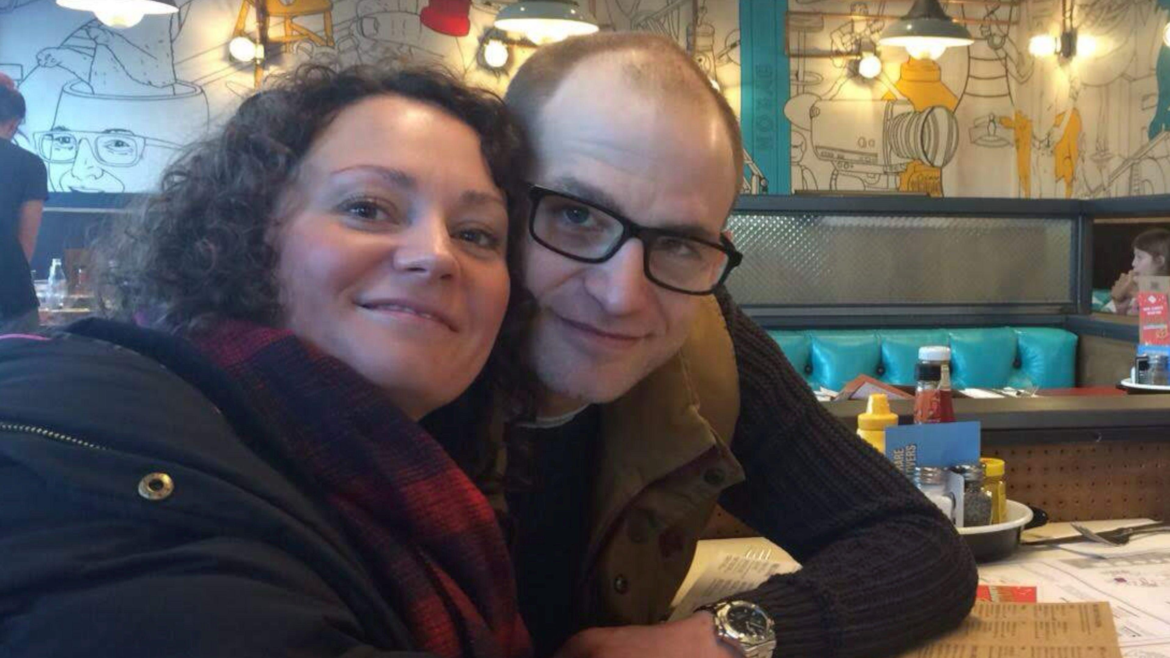 Emma Harley sits next to her brother Terry White at a table in a restaurant. Both are wearing dark tops. Emma has dark curly hair and wears a dark blue and red scarf. Terry wears dark rimmed glasses and a silver wristwatch. Both are smiling at the camera