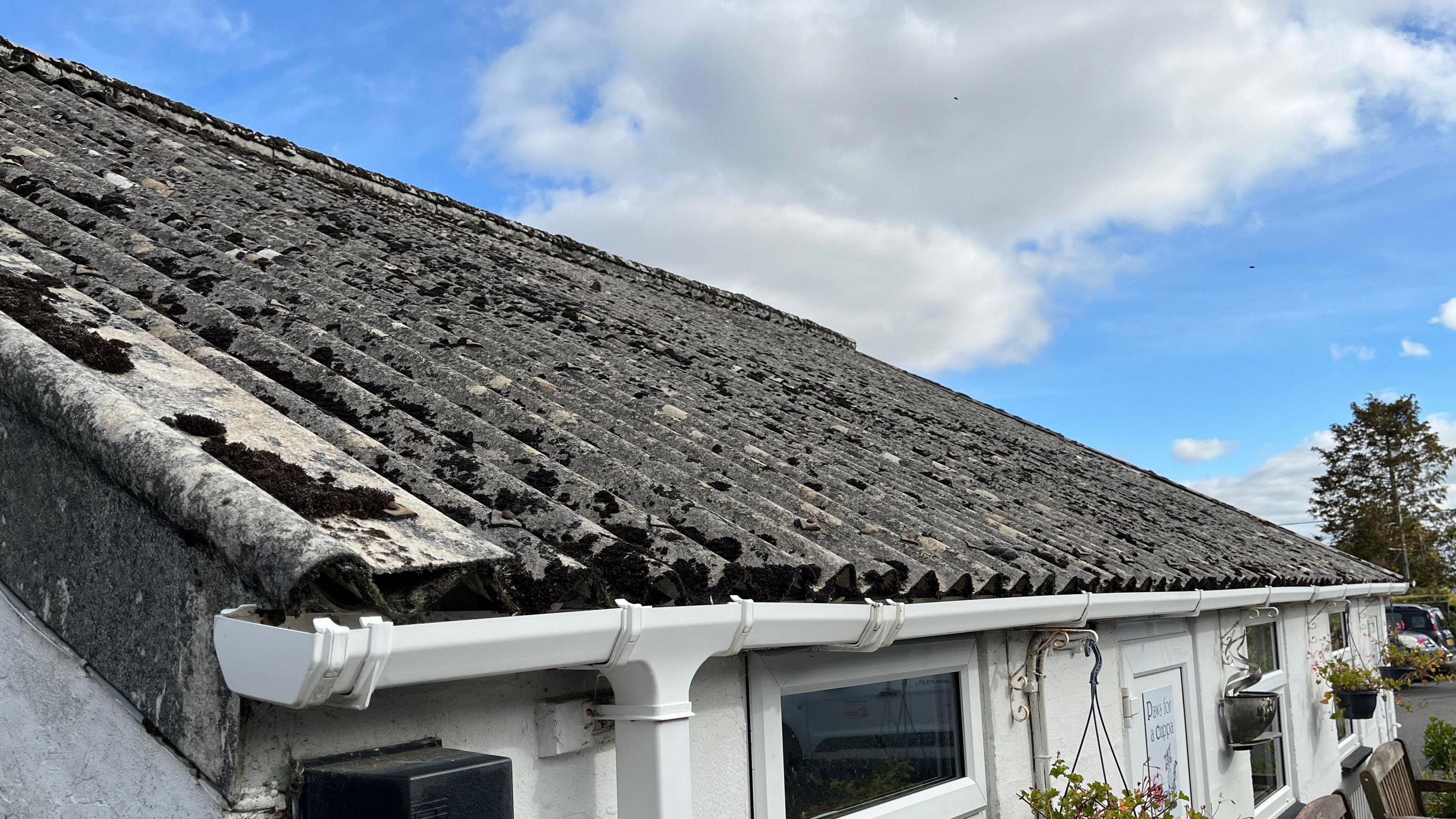 A single-storey white building with a grey pitched roof. The roof is covered in corrugated iron, which is covered in lichen and moss. The sky is blue with a few clouds. 