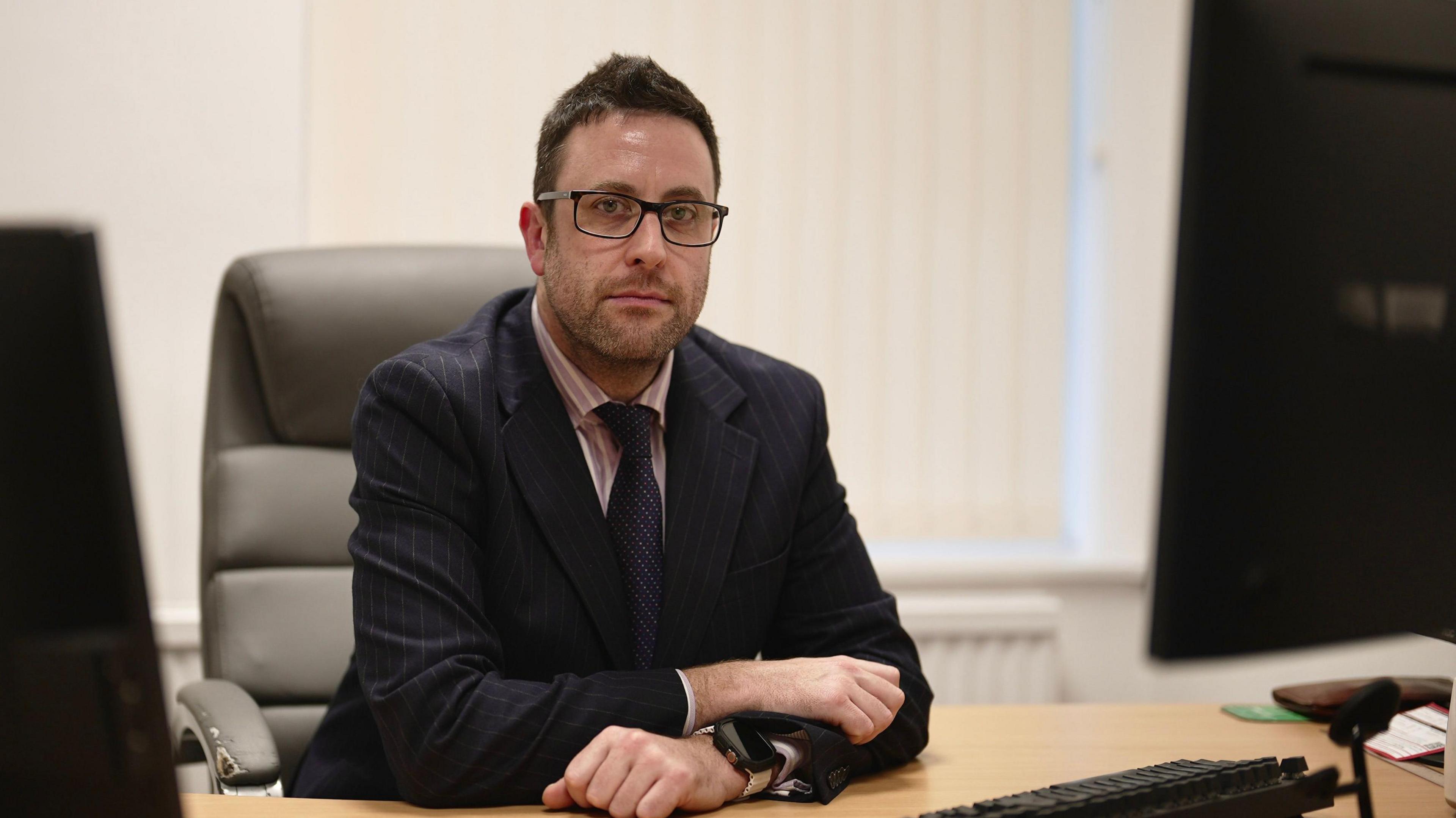 Damien Dobson, a man with short, dark hair, neatly trimmed stubble and dark-framed glasses, wearing a pinstriped suit and a striped shirt, sitting at a desk in his office on a leather chair.