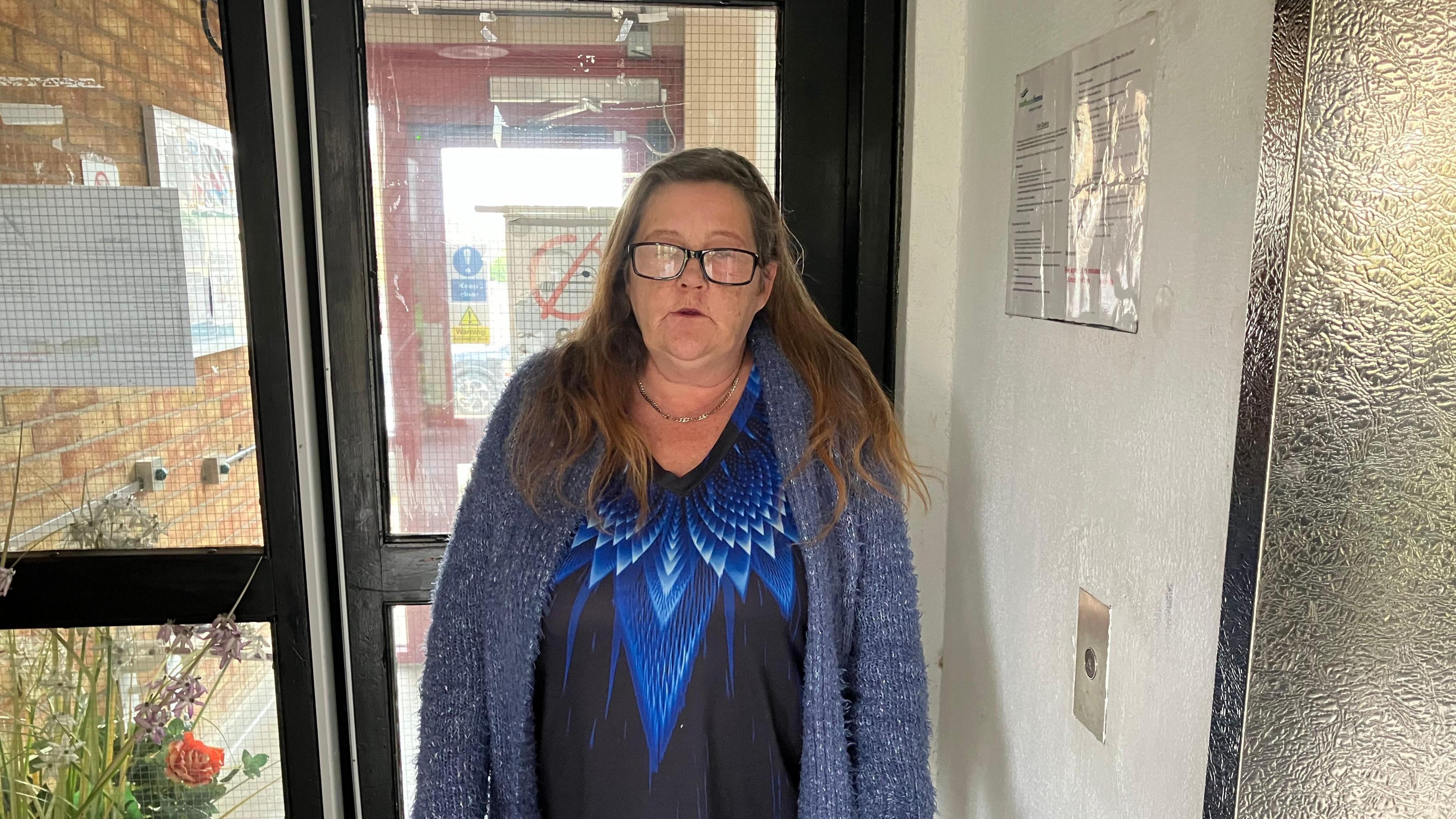 Zeta is wearing a blue cardigan and a dark blue patterned top underneath. She is standing next to a lift at Yantlet House.