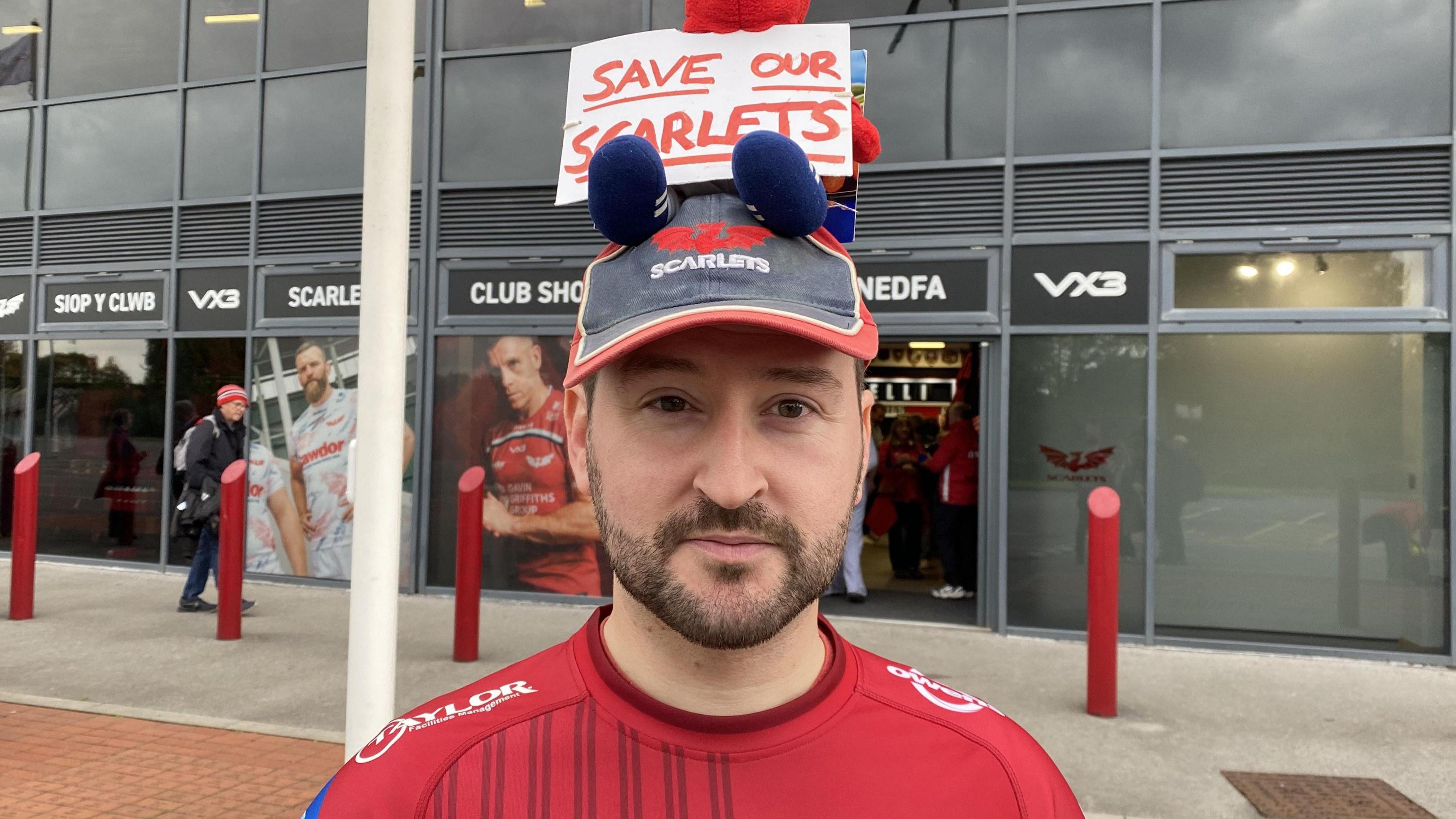 Mae Tomos Davies yn gwisgo crys rygbi a chap Scarlets.