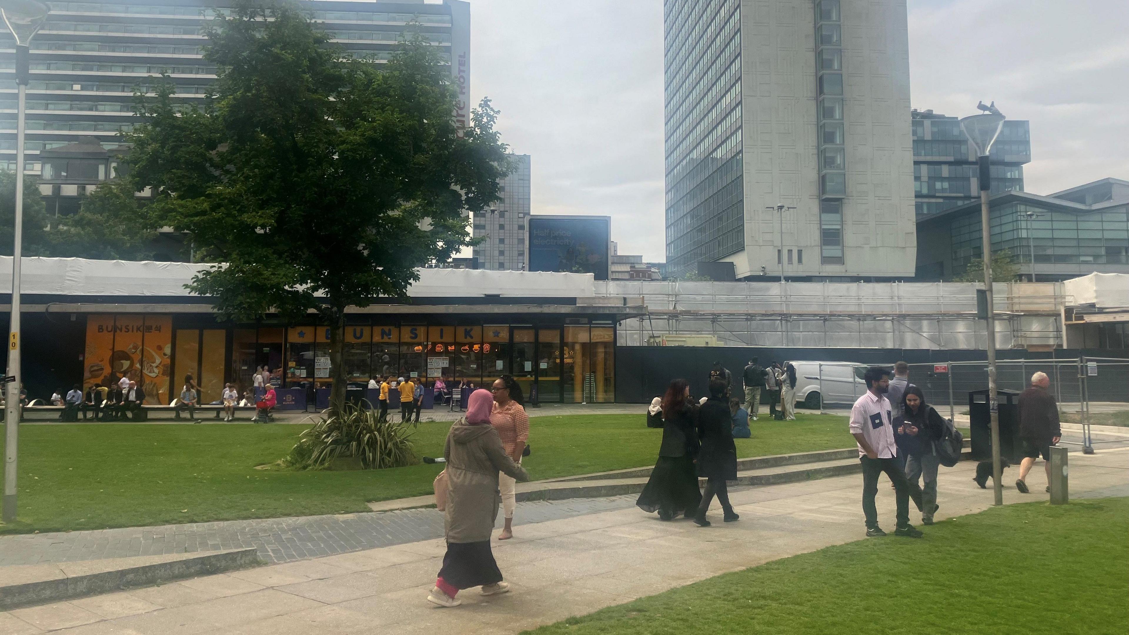 Pedestrians walk along a flagged path through a grassed area in Piccadilly Gardens in Manchester. Tall office blocks can be seen in the background. 