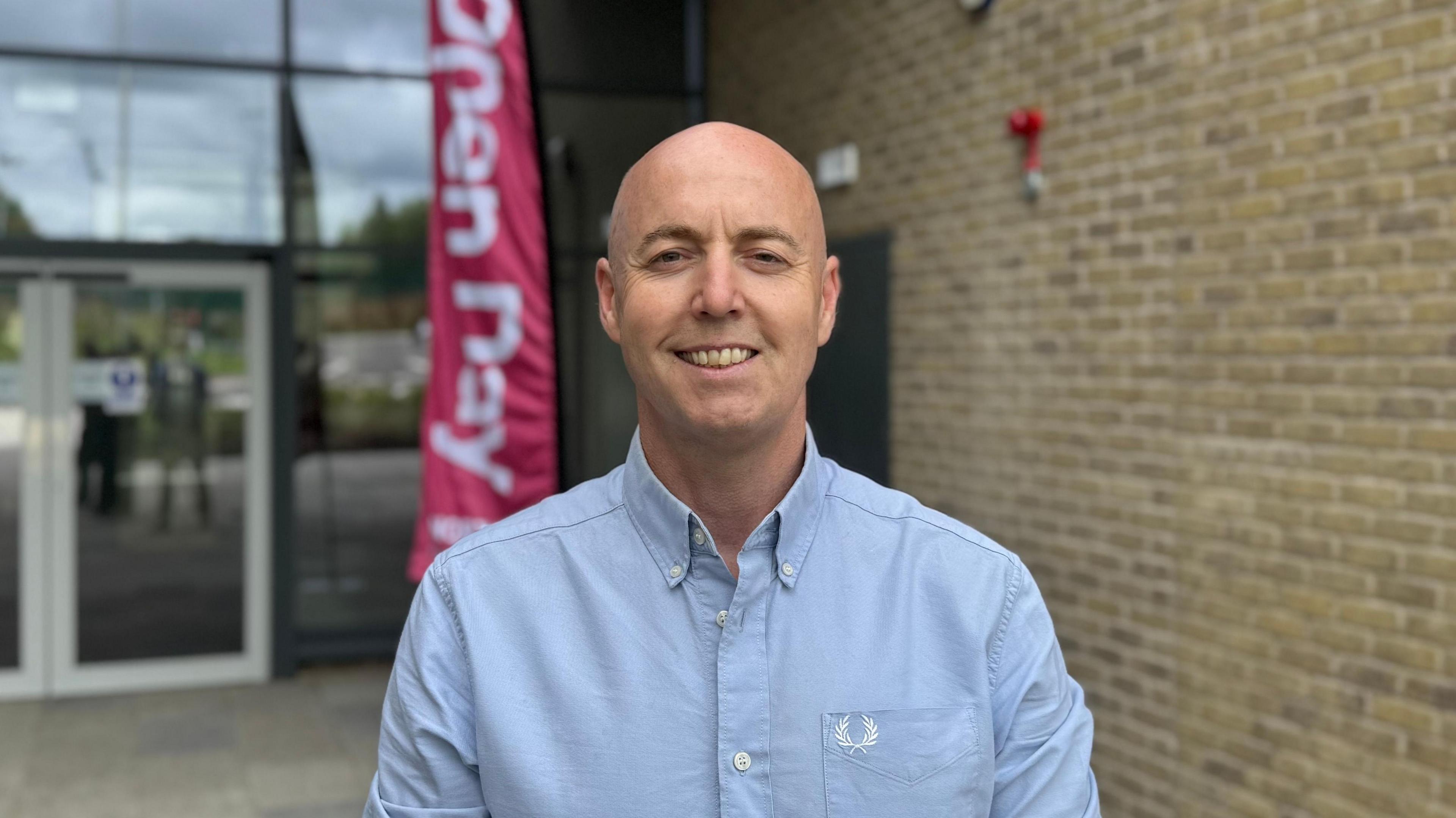 Jon Burrows is a middle-aged man with a shaved head. He is wearing a light blue shirt with white buttons. He is smiling.