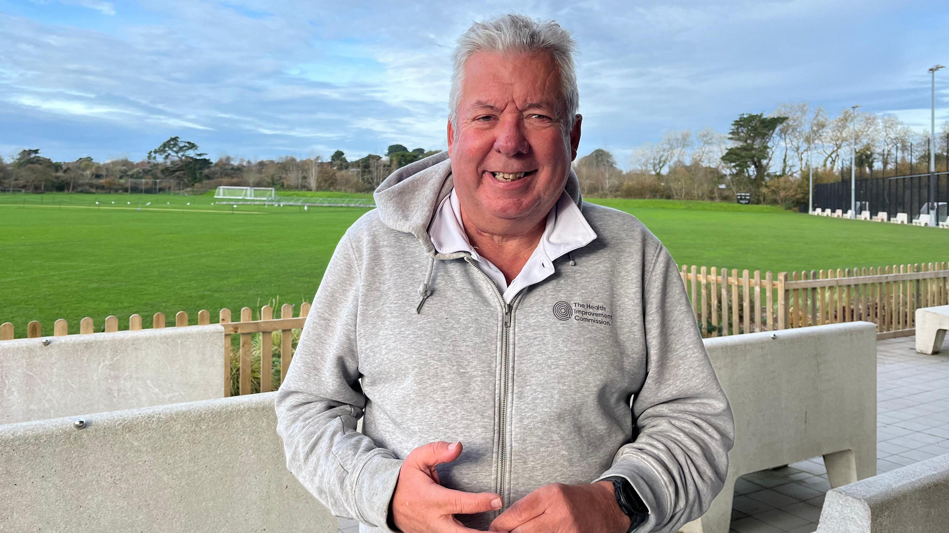 Alun Williams, Be Active Lead at The Health Improvement Commission, standing at the KGV playing fields. The sky is very blue and the grass is well-trimmed and green. There is wooden fencing and a few football goals in the distance. 