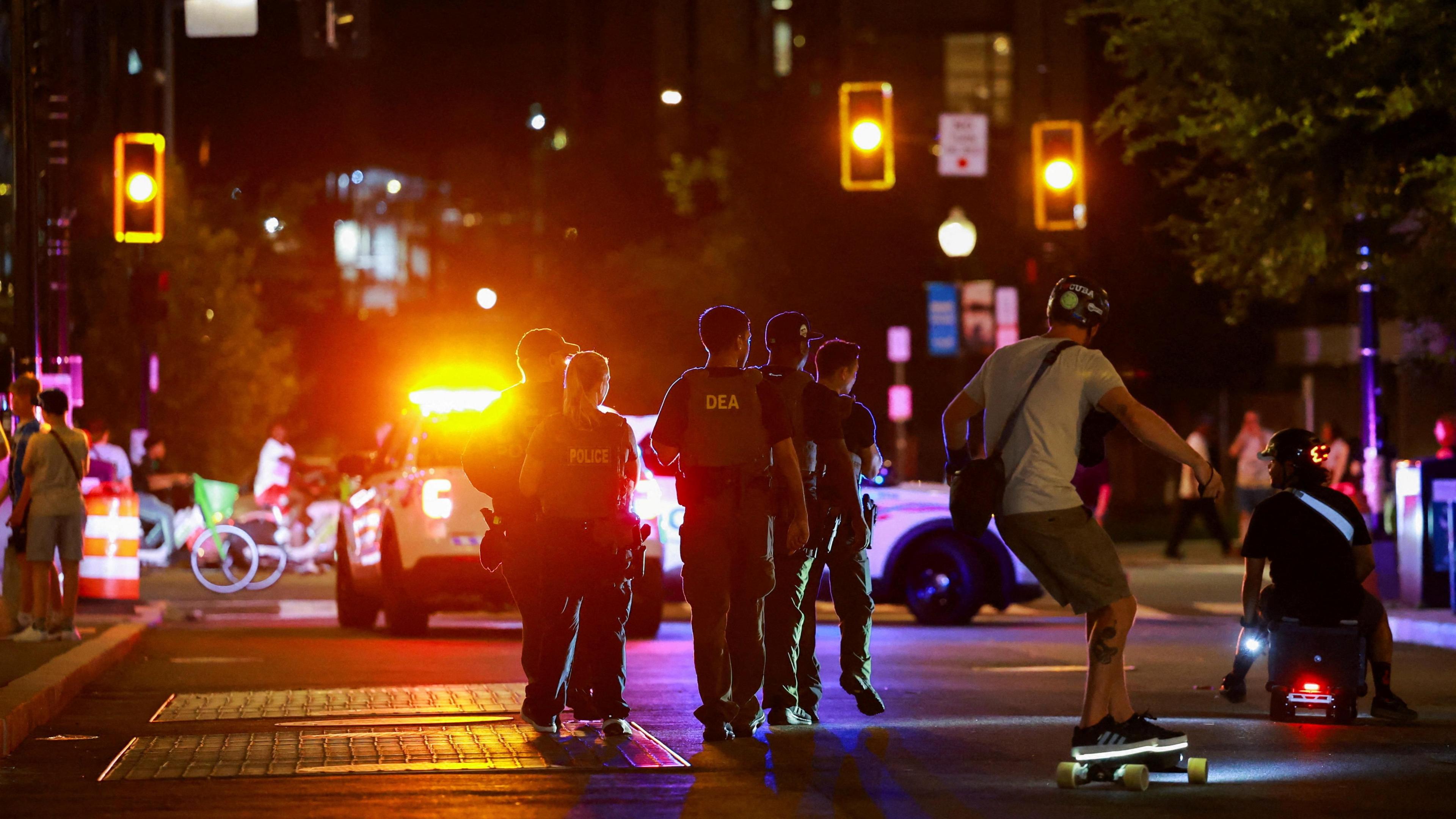 A street at night with glowing traffic lights and pedestrians standing around, as a truck stops in the background and five uniformed agents walk in the middle of the street, where a man also rides a light up skateboard and another sits on some sort of wheeled toy