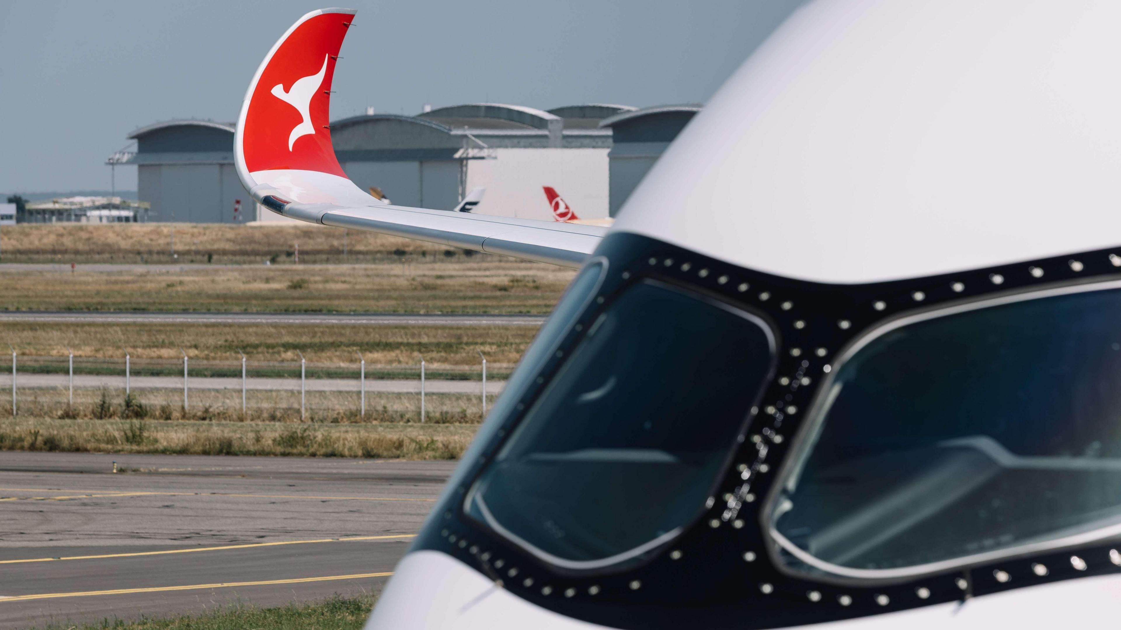 A close up of an airplane's cockpit, with the wingtip visible in the distance with the red and white kangaroo logo of Qantas.