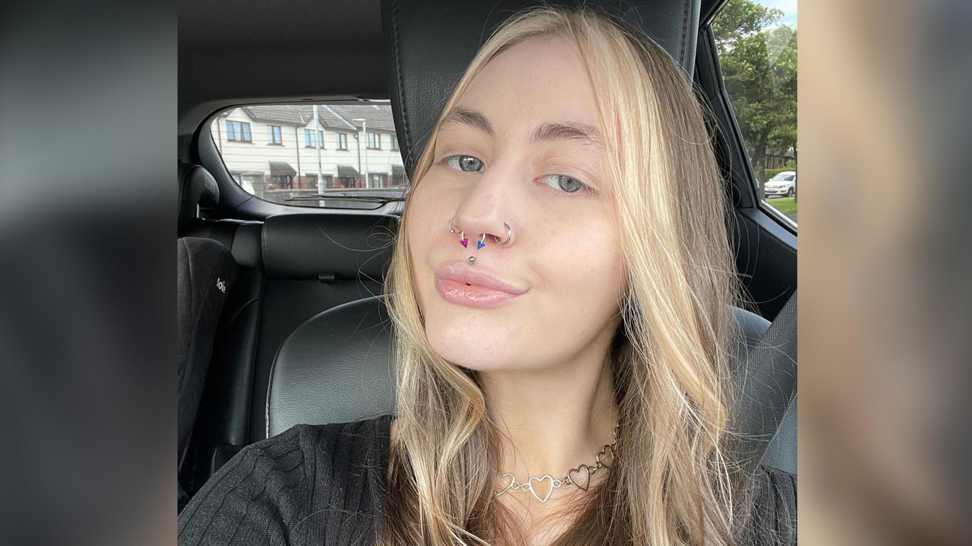 A selfie Kelly has taken in a car with black leather seats. Her seatbelt is covered by long, wavy brunette hair which has blonde dye through it. She is wearing a black jumper with a striped pattern. A silver necklace made of a string of heart shapes rests on her neck. Kelly, who has green eyes, is pouting and has multiple piercings, including a silver stud in the centre of her upper lip. She has two silver hoops on her outer nostrils and a septum piercing, which has two pyramid shaped ends, one coloured pink and the other blue.  