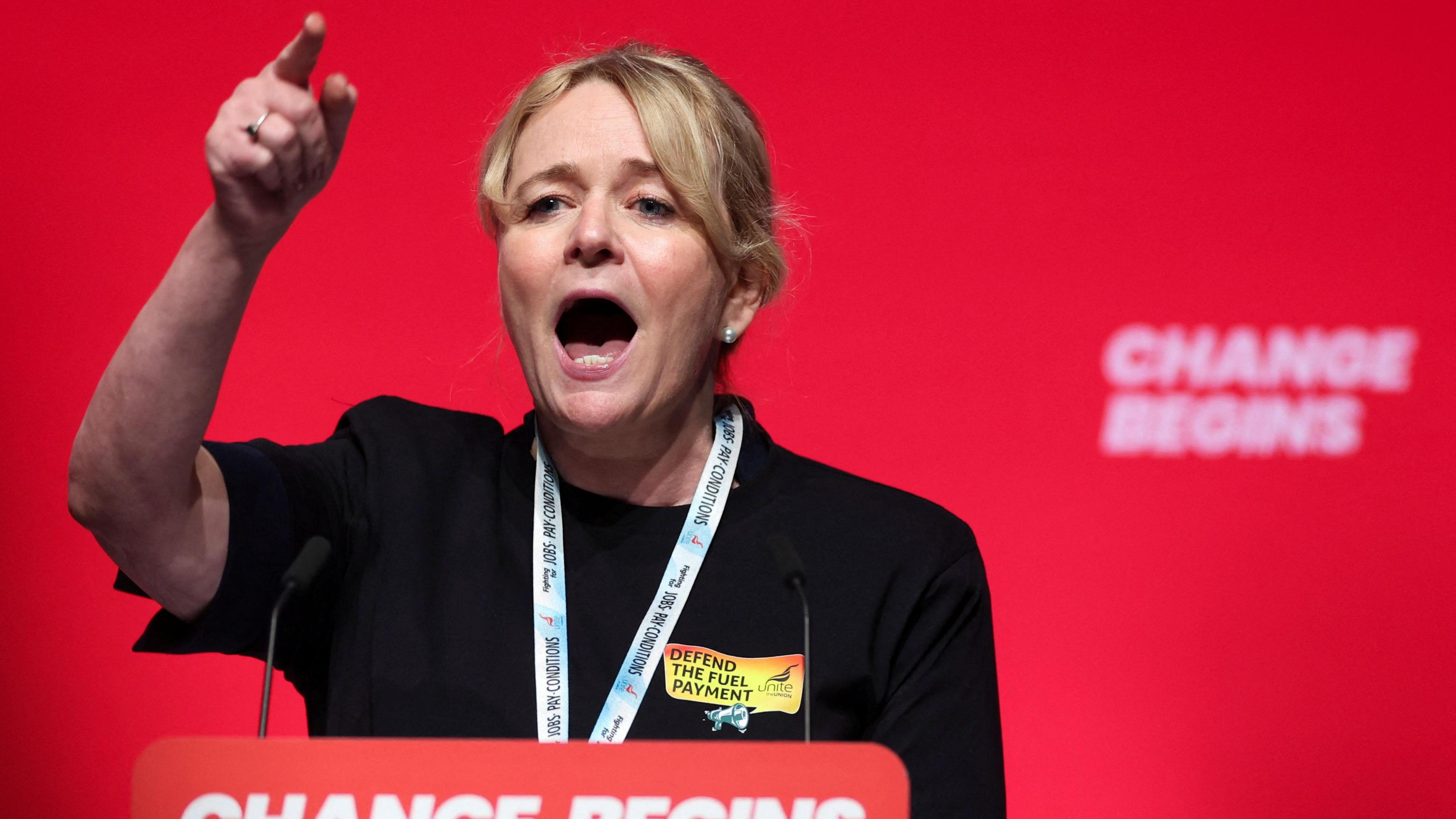 Sharon Graham - a woman with blonde hair and wearing a lanyard and black outfit - stands in front of a red wall behind a podium with a microphone, pointing in the air as she speaks.