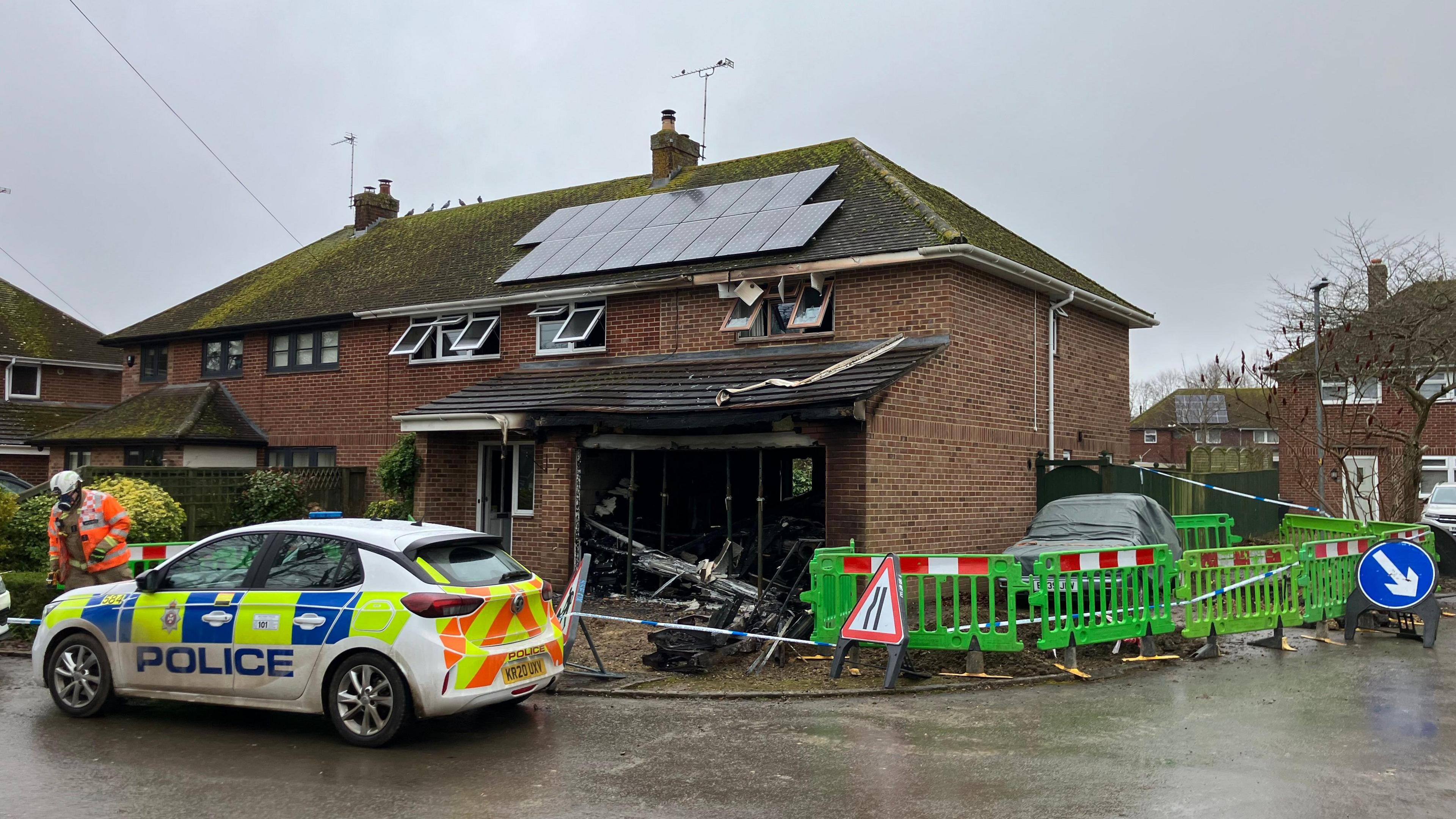 A house down Ferozeshah Road in Devizes. It is red brick and has solar panels on the roof. It has been damaged by fire - all the windows are open and there is soot on the tiles. Debris can be seen in front of the house. There is a green fence cordoning off the side of the house, as well as police tape and a blue road arrow telling drivers to pass on the right. A police car is parked outside and a man can be seen in an orange high vis jacket near it. 