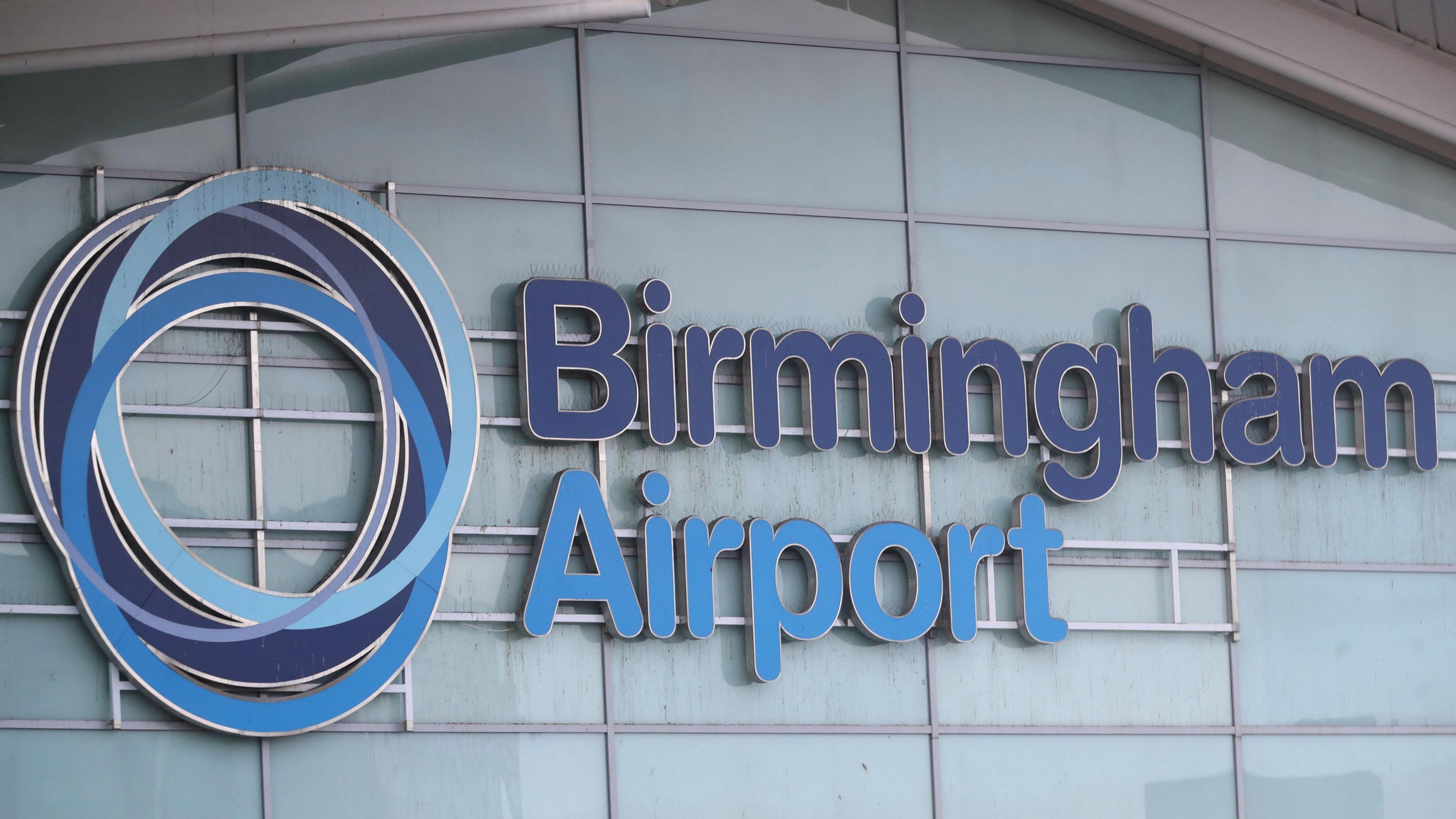 A close-up photo showing the windows of a building, with a sign across the windows that says "Birmingham Airport" in blue letters along with a logo made up of blue circles.