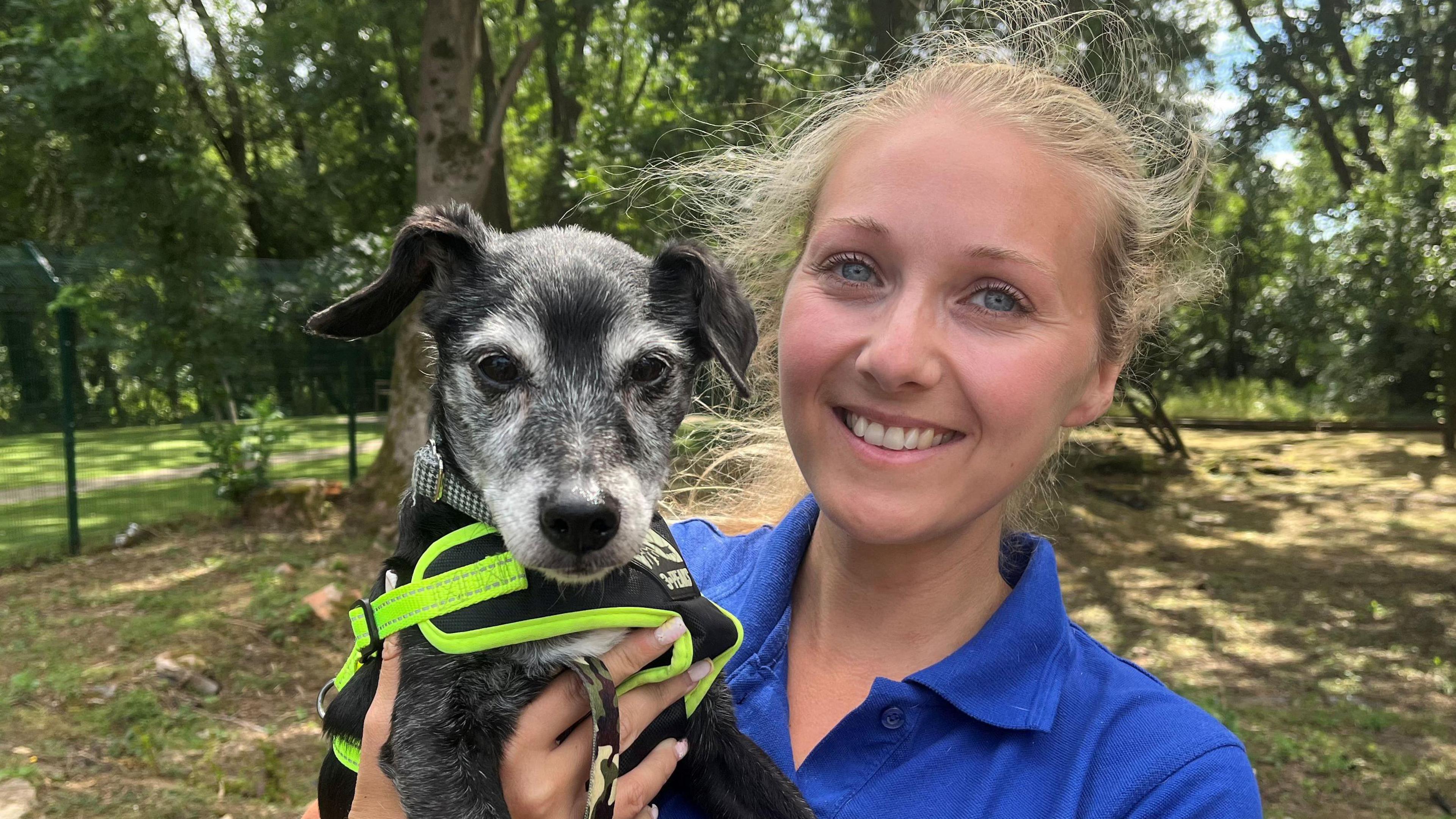 A woman with tied back blonde hair and blue eyes smiles at the camera, wearing a blue T-shirt. She is holding a terrier puppy to her left, who wears a neon harness that says '3 peaks'. The dog looks at the camera. They are standing on patches of grass with trees behind them and a green metal fence to the left. Sunlight is dappled through the shadows of trees behind them.