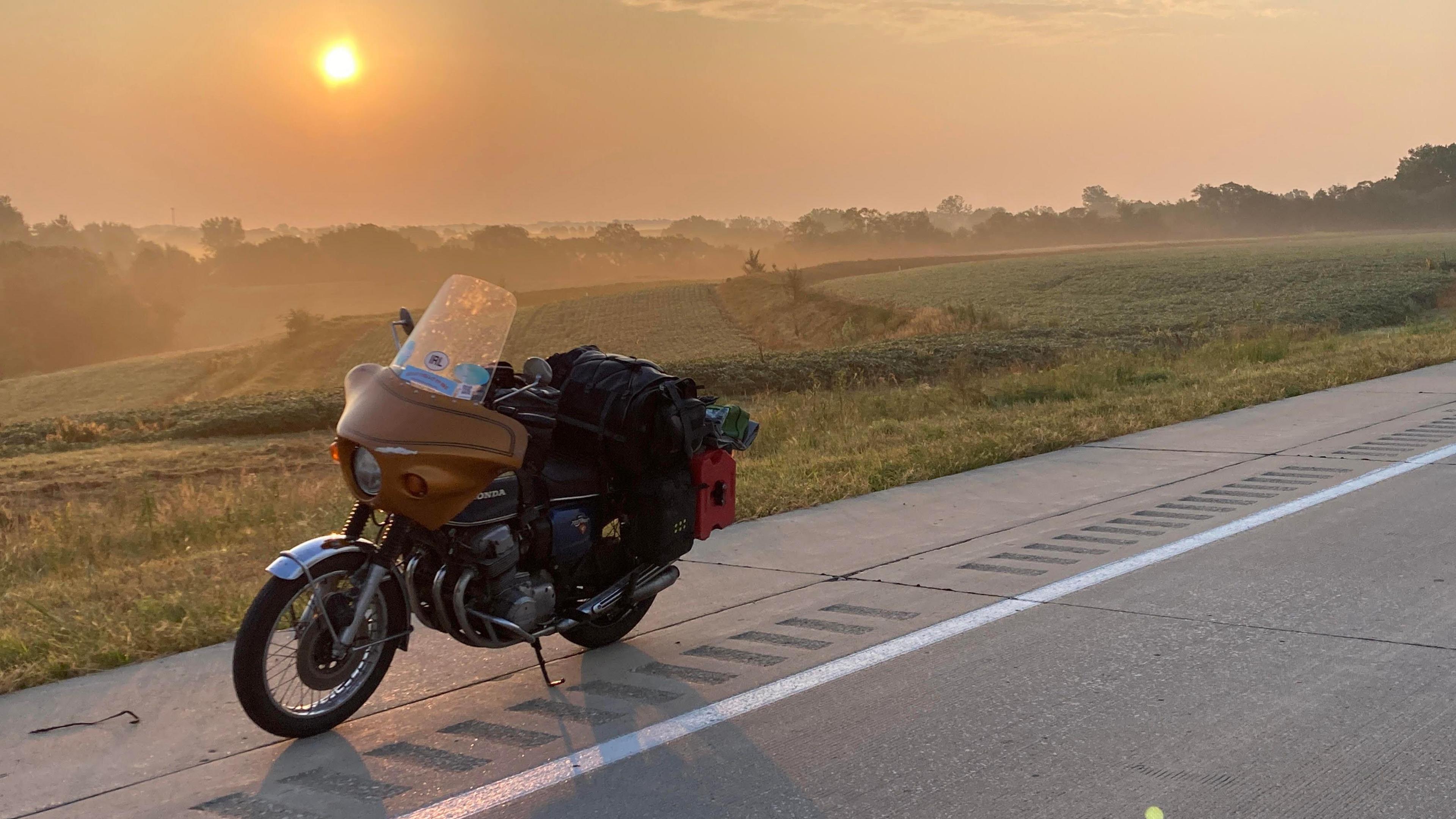 A motorbike is parked by the side of a country road in America with sun beginning to set in the distance amid rolling farmland