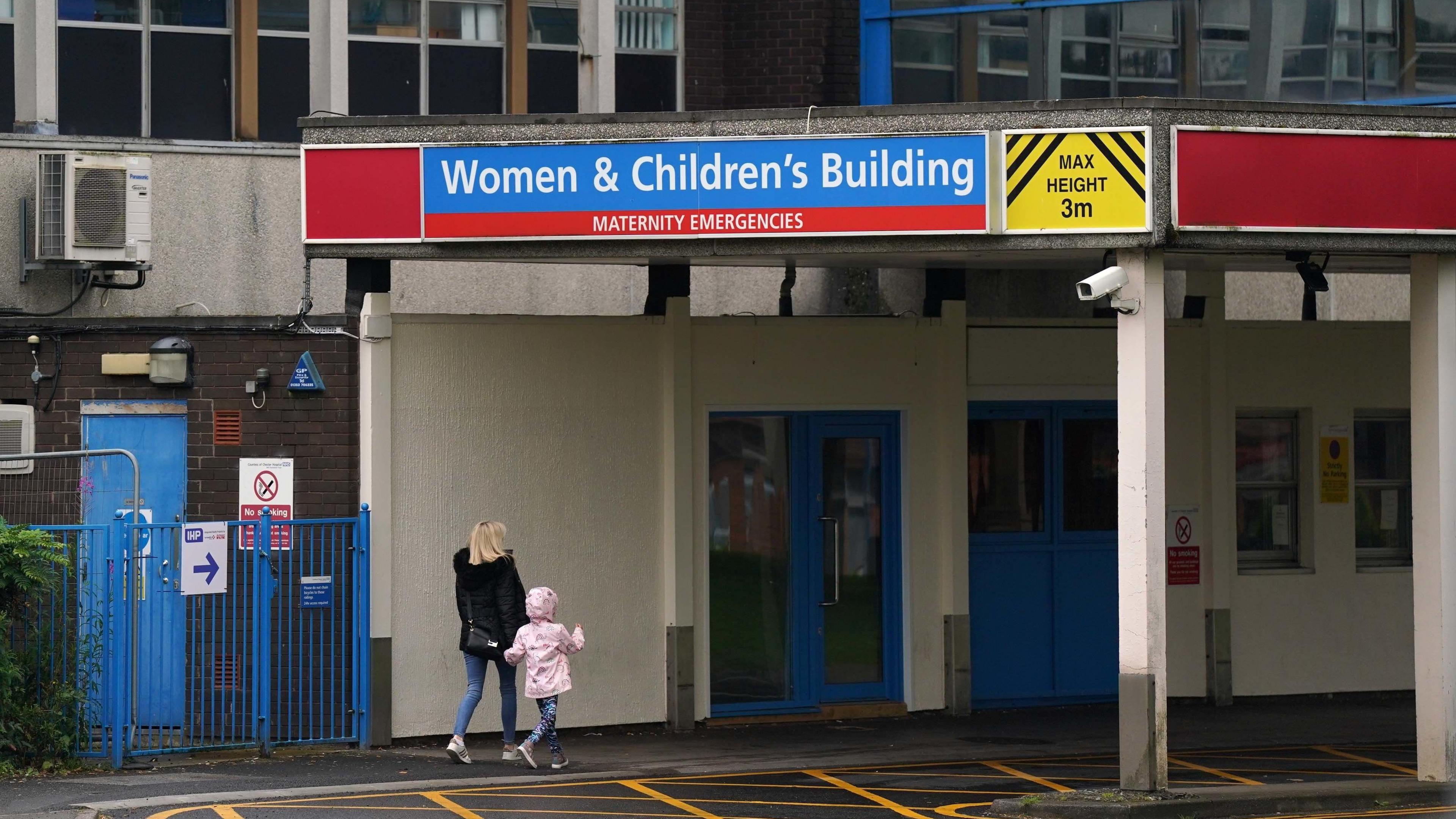 A blonde woman in a black coat and blue jeans walks into the Women & Childen's Building at the Countess of Chester Hospital holding hands with a young girl, who has a pink coat with the hood up and blue leggings. 