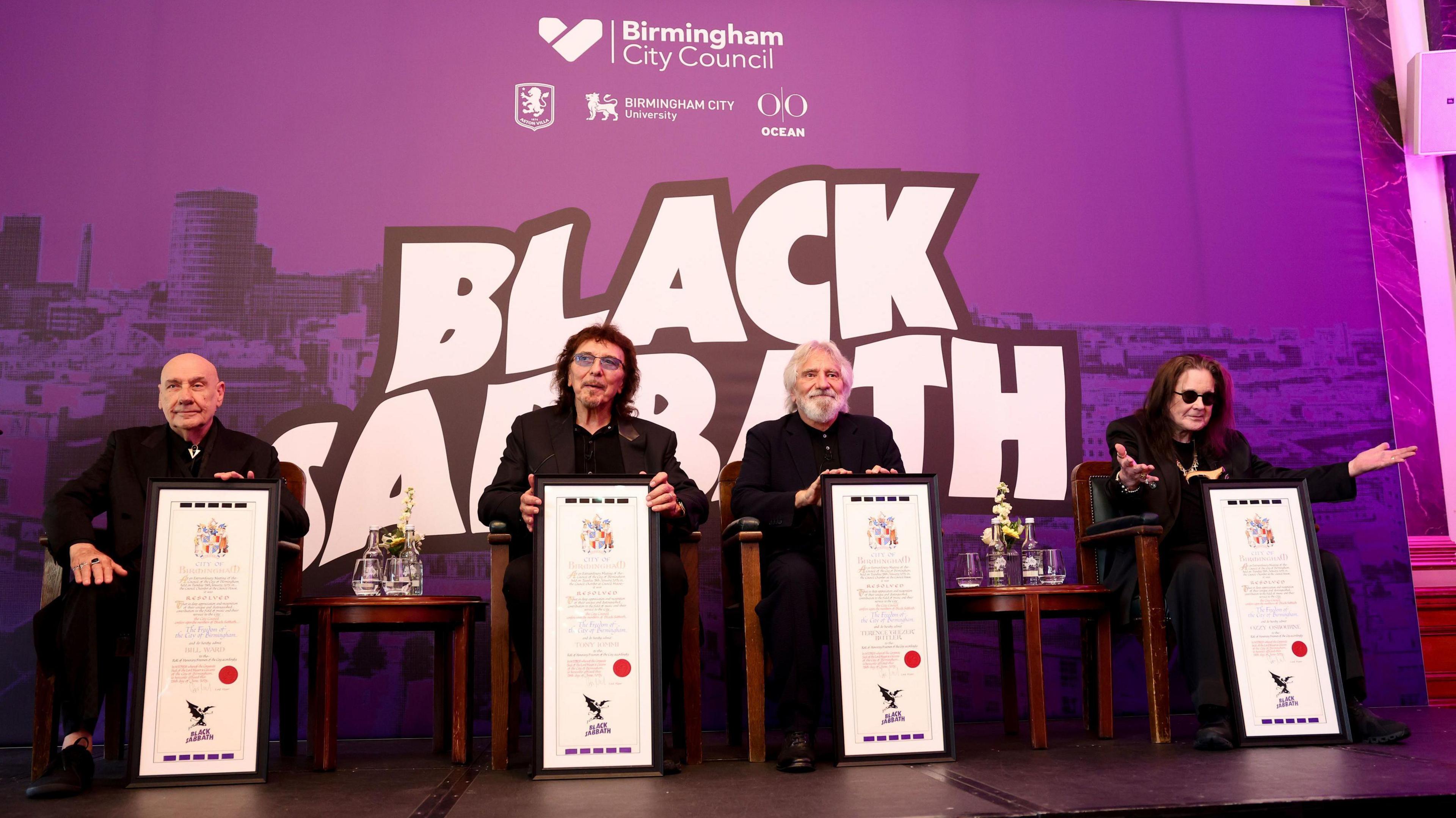 Bill Ward, Tony Iomi, Geezer Butler and Ozzy Osbourne sit behind framed certificates. A purple Birmingham City Council backdrop has the name Black Sabbath in white on it. 