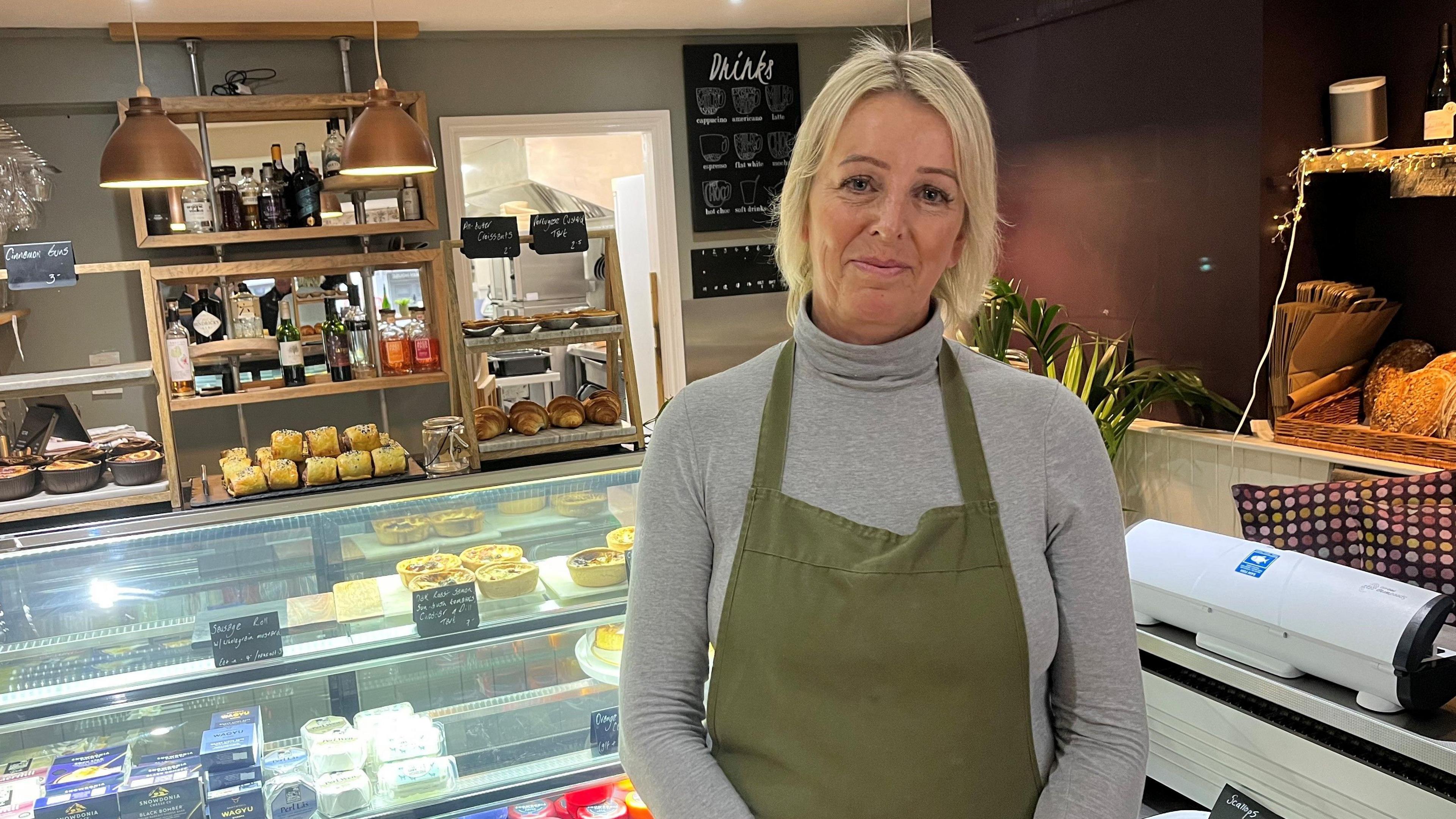 Emma Downey has short blonde hair and is wearing a grey roll neck jumper and green apron. She is stood in the shop front of a deli