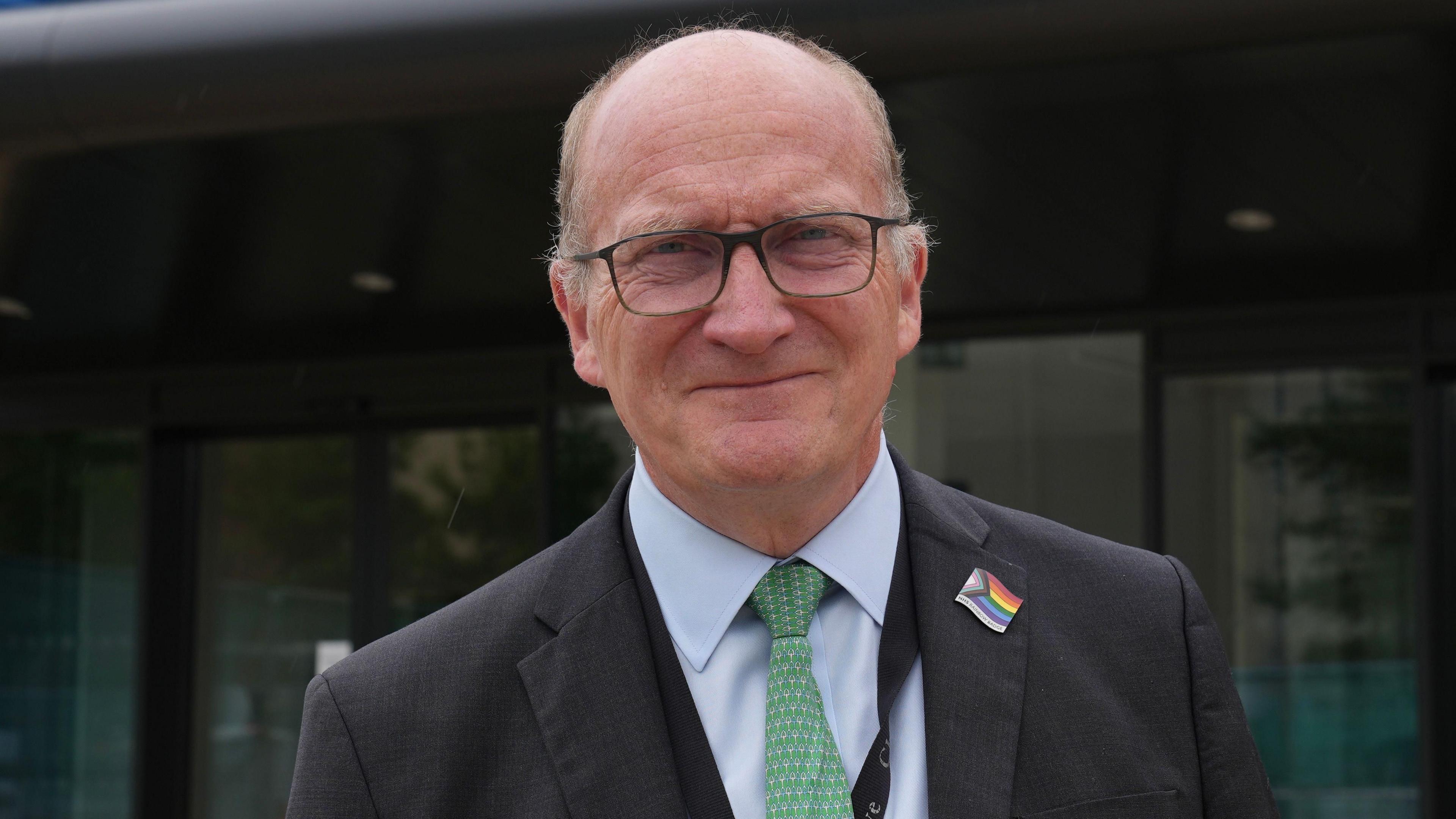 Nick Hulme in a black suit jacket with a blue shirt and a green tie. He is wearing glasses and is looking into the camera and smiling.