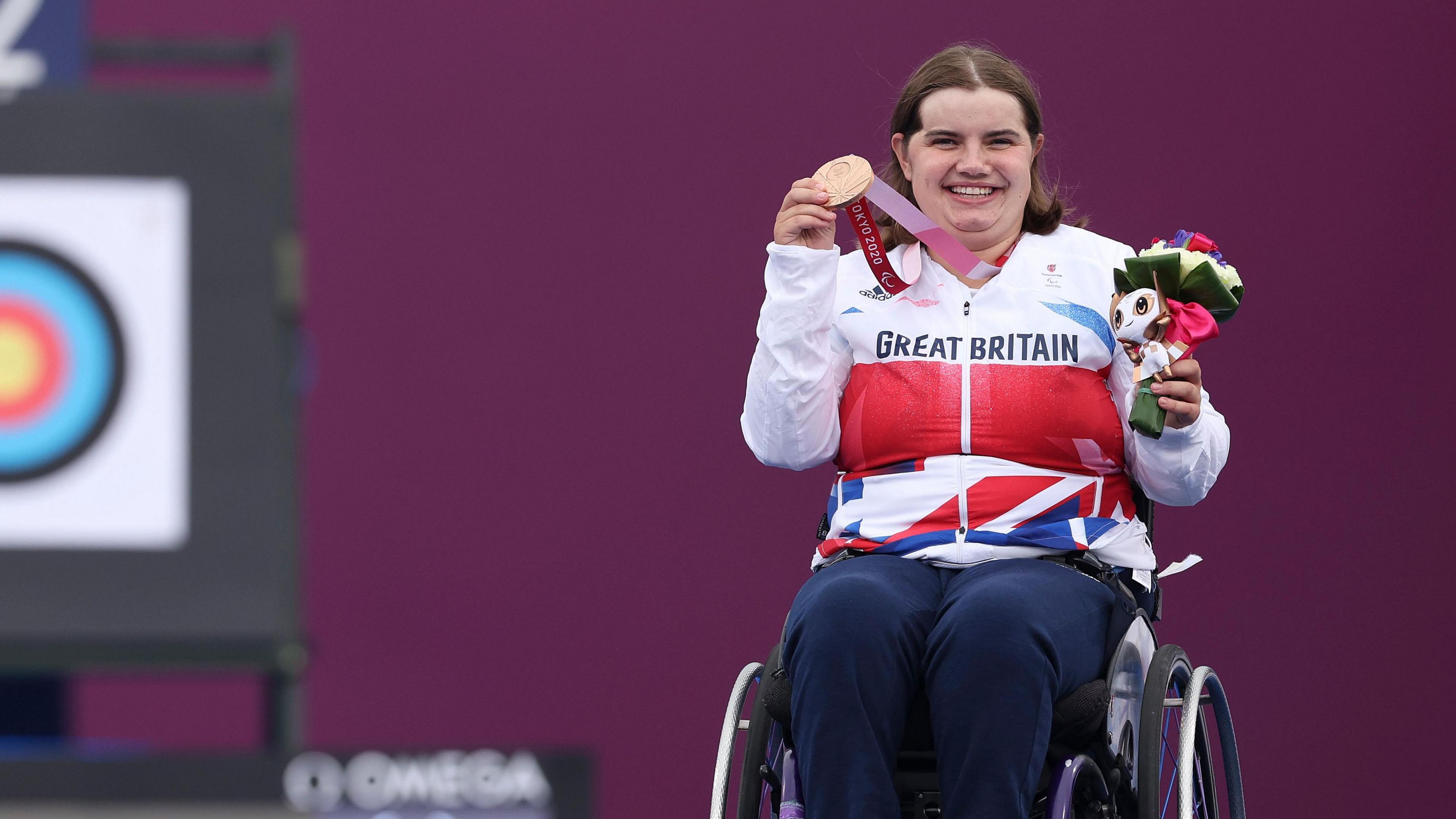 Bronze medallist Victoria Rumary of Team Great Britain poses during a medal ceremony on Day 8 of the Tokyo 2020 Paralympic Games in Tokyo. She is wearing her white, red and blue GB kit, and is holding up her bronze medal with her right hand and holding a small bunch of flowers in her left. She is in a wheelchair. An archery target can be seen in the background.