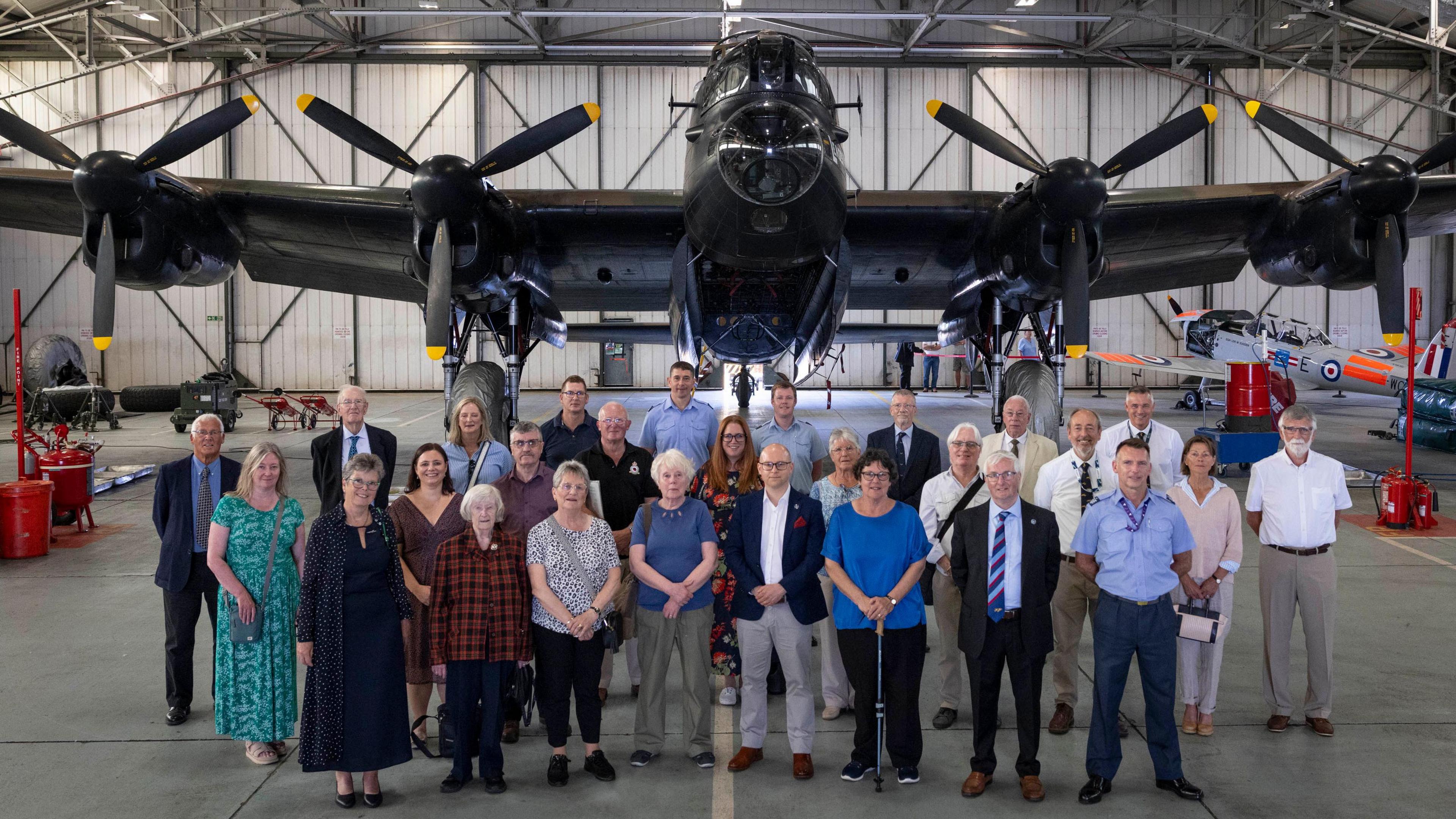Group of people standing in front of a Lancaster bomber housed inside a hangar. They are all looking towards the camera.