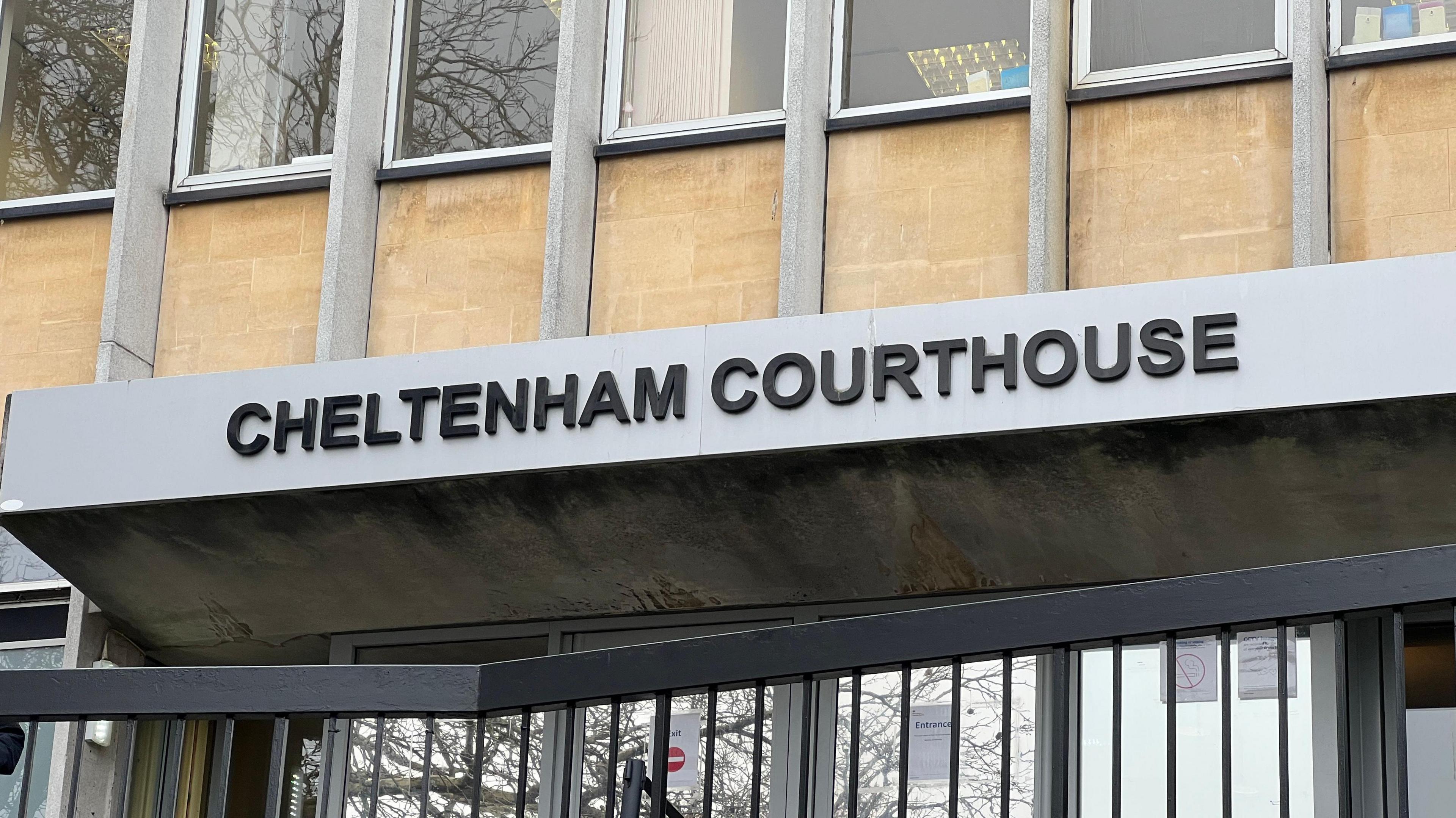 A close-up shot of the entrance at Cheltenham Magistrates' Court. The building is beige with grey panels either side of the windows. "Cheltenham Courthouse" is written above the entrance in capital letters