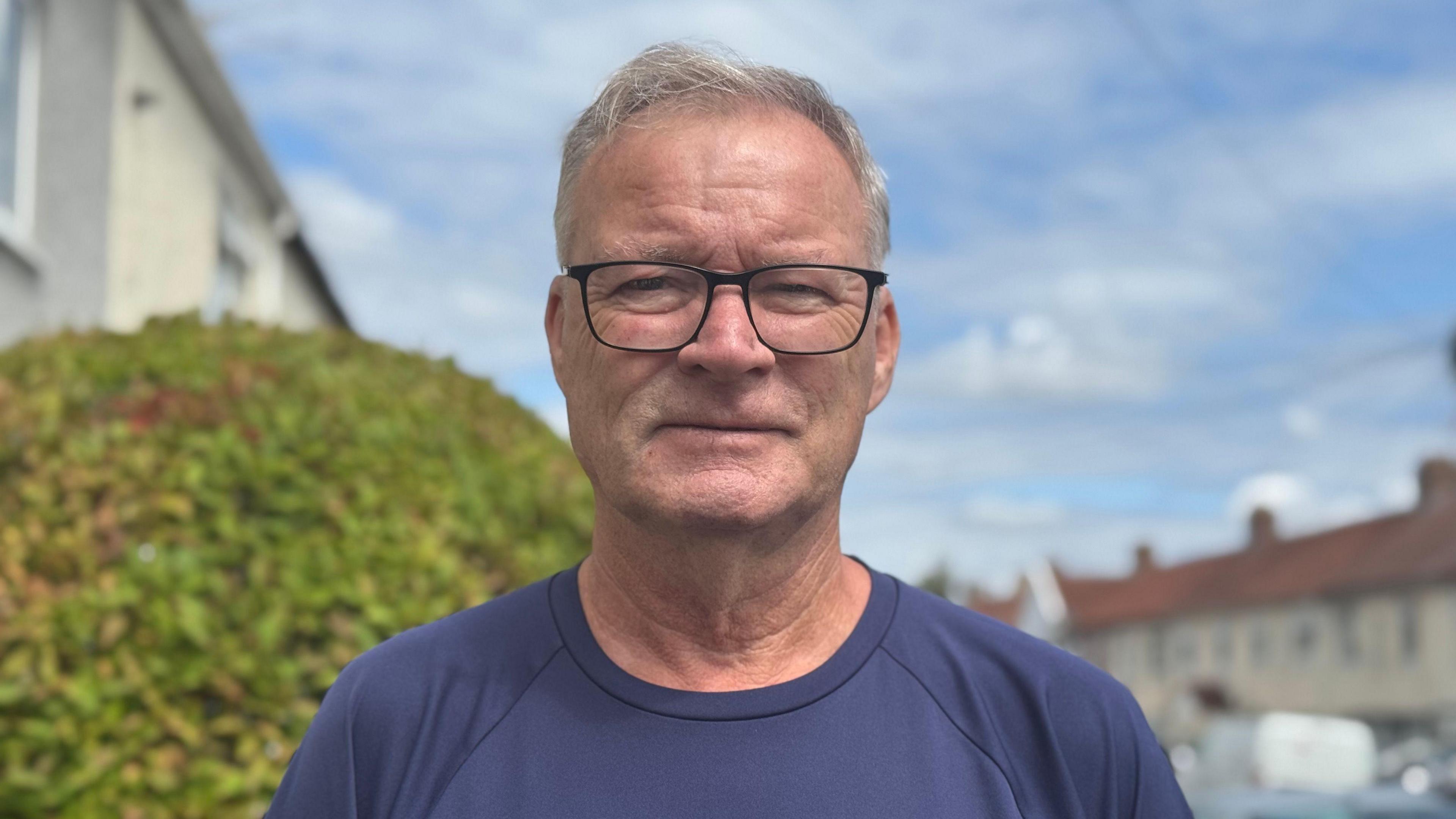 Kevan Feltham looks at the camera in his front garden, he wears black rimmed glasses and has short, grey hair. He is wearing a navy blue T-shirt. There is a hedge behind him.