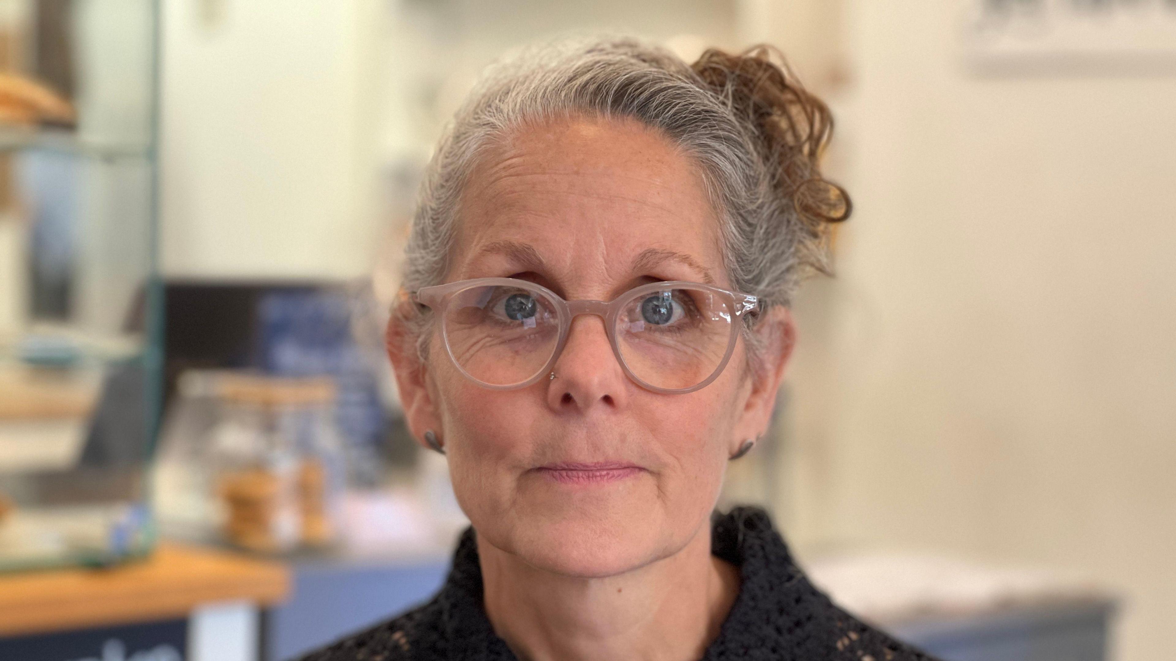 Image shows a lady sat in front of a shop counter.
She has glasses and her grey hair is tied up in a bun. She is wearing earrings and a black cardigan. 