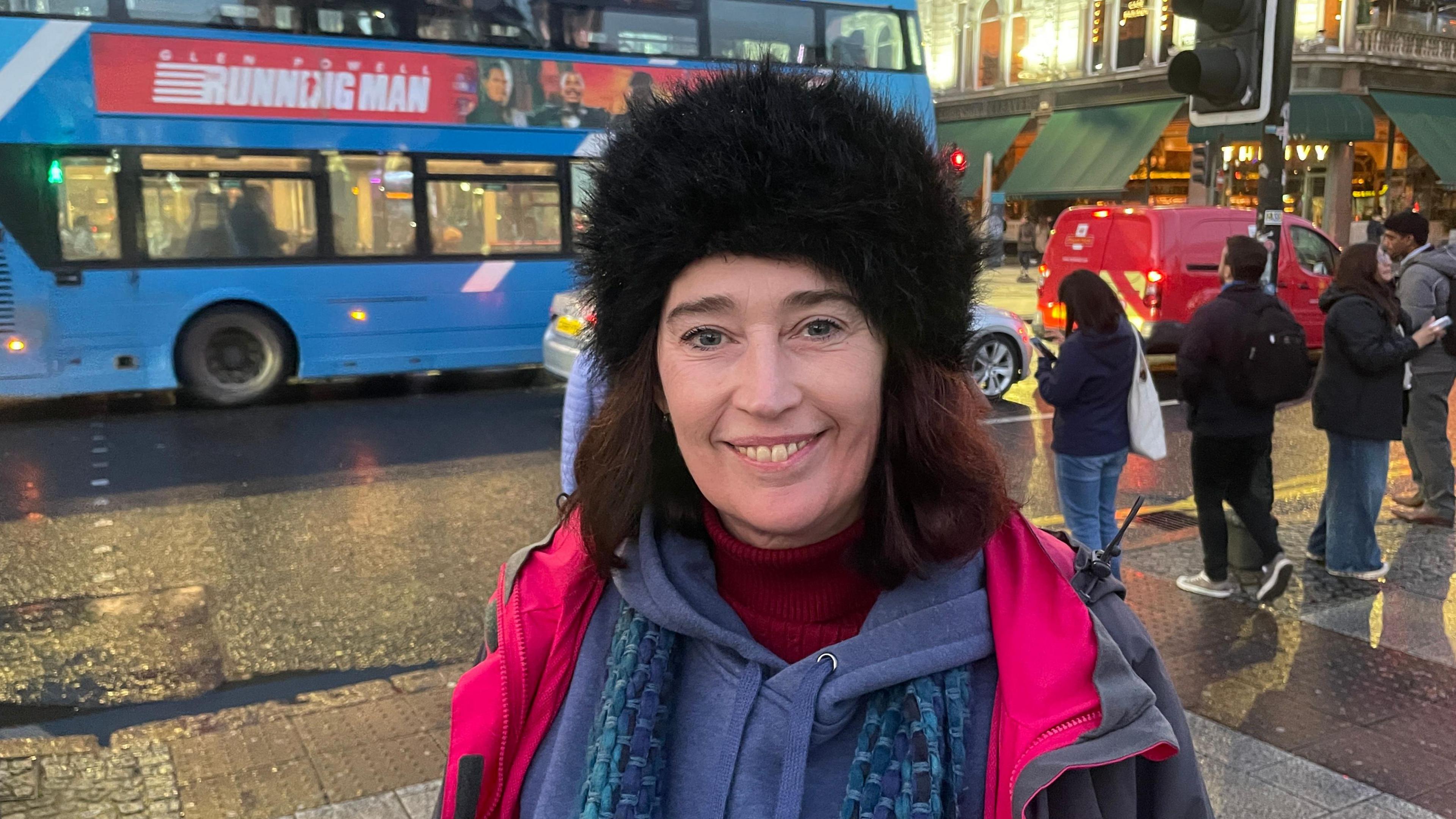 Anna Bettess wears a black furry headband and has brown hair. She is wearing a blue hoodie and a grey and pink jacket. She is standing outside and there are people and a bus behind her.