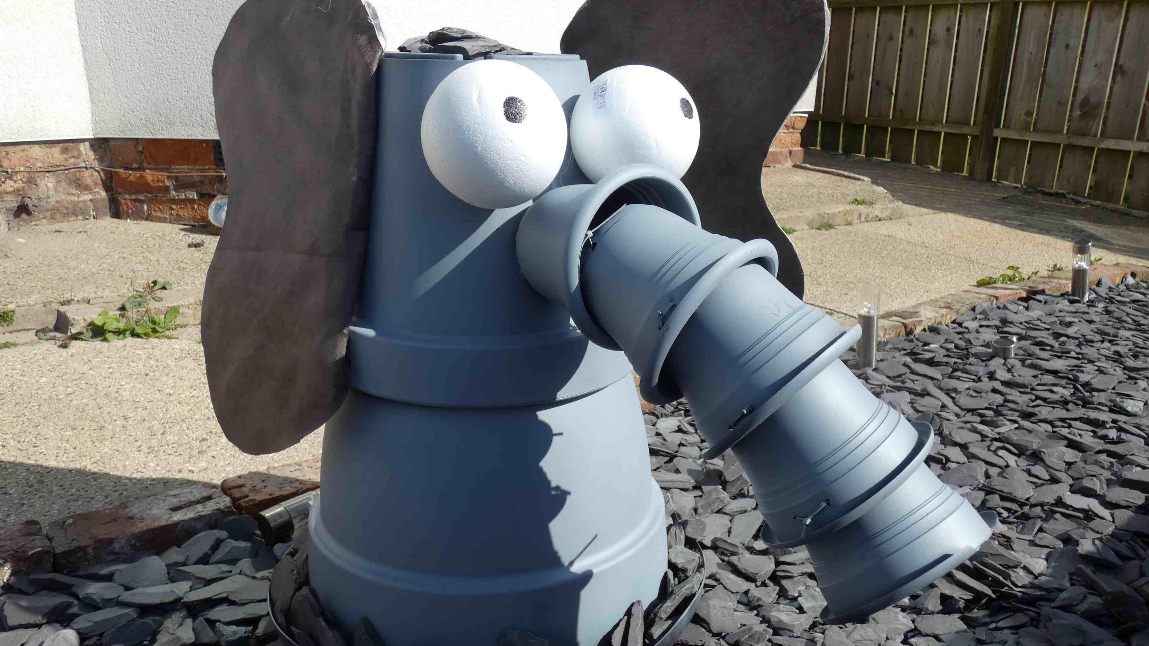 A grey elephant's head made out of plant pots. It is on grey slate in a garden with concrete and a few weeds behind it, a white wall and a wooden fence.