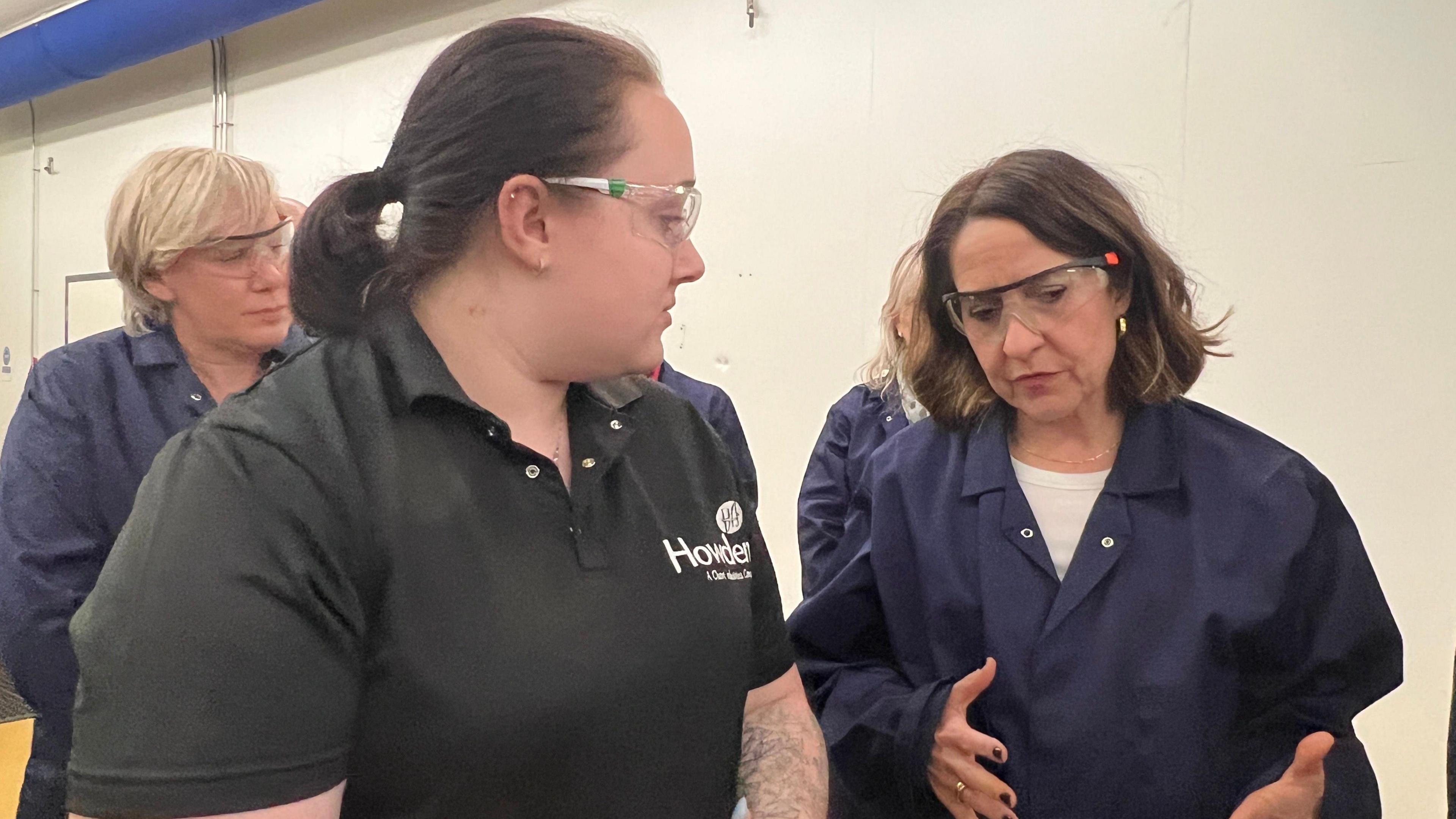 Liz Kendall is wearing dark blue overalls and safety glasses, she is speaking with a woman wearing a black polo shirt and safety glasses, behind her are several people also wearing overalls, they are in a room with white walls.