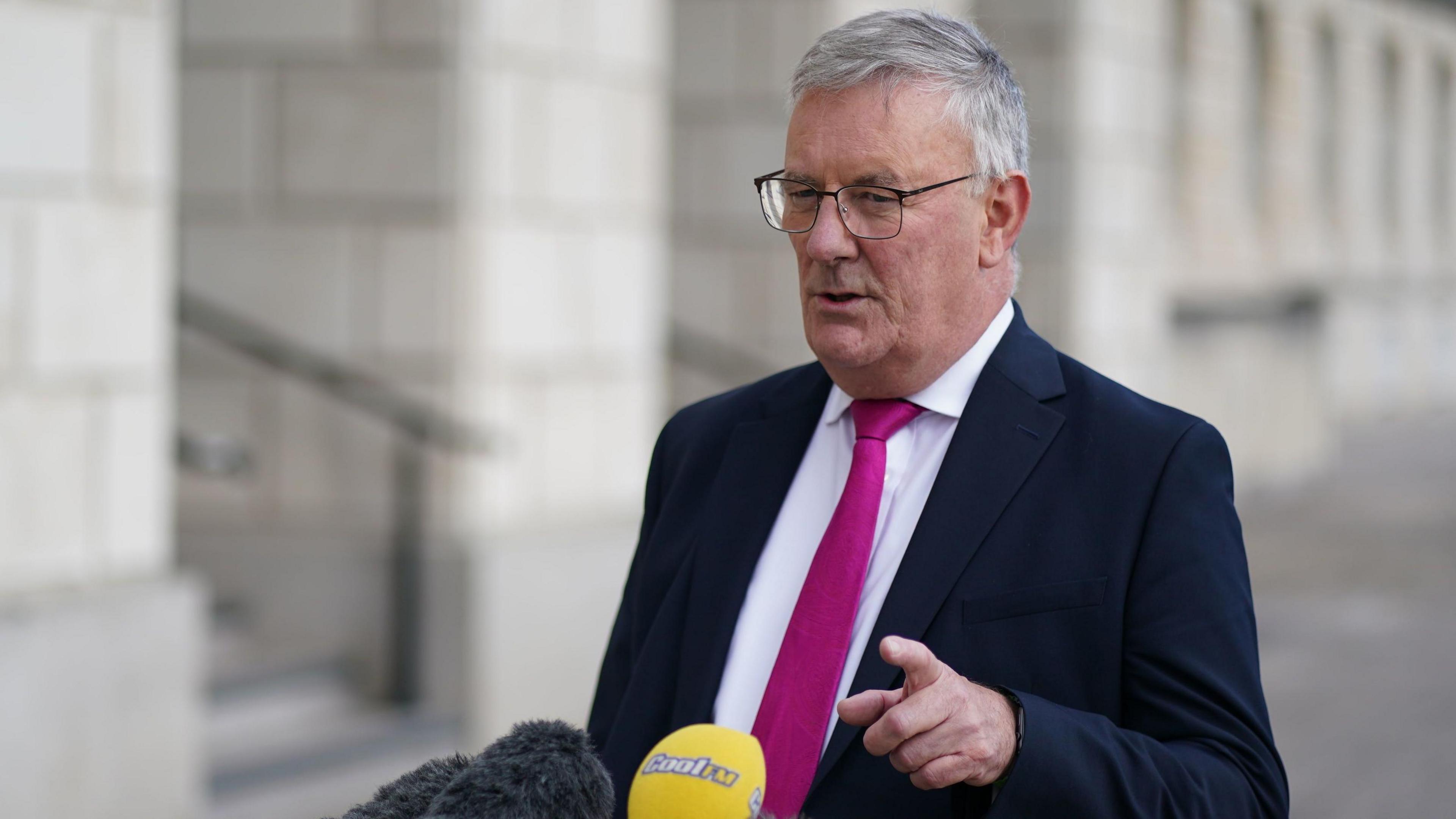 A man with white hair and dark rimmed glasses talks to someone off to the side of the camera while pointing. He's standing in front of a number of grey and yellow microphones. He is wearing a navy blue suit, a white shirt and a bright pink tie. The background is blurred, but you can make out light grey stone walls and steps with black railings. 