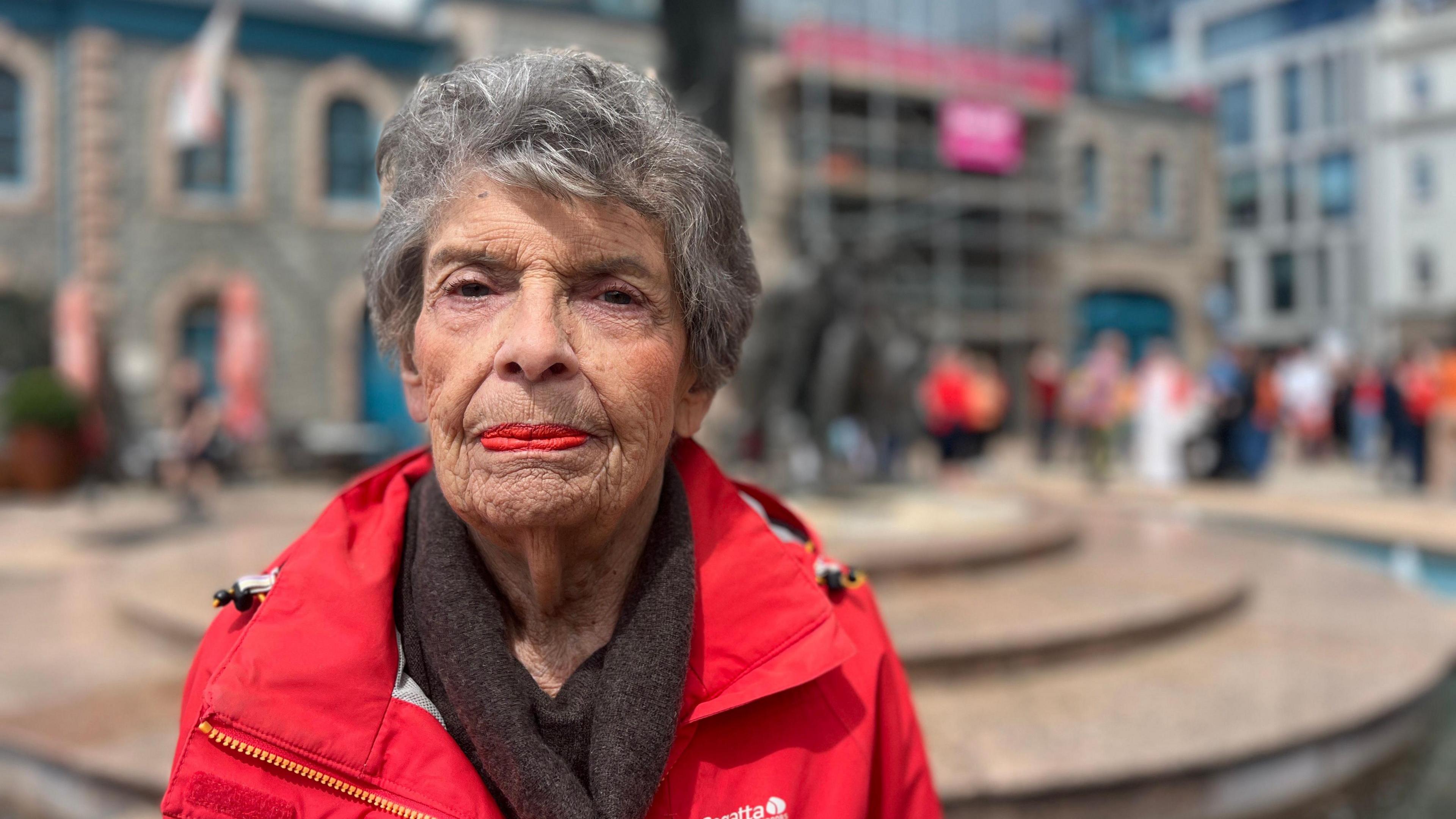 Susan looks at the camera as people in orange gather behind her and there's a statue behind her. She's wearing a red coat and a dark brown scarf and has short grey hair.