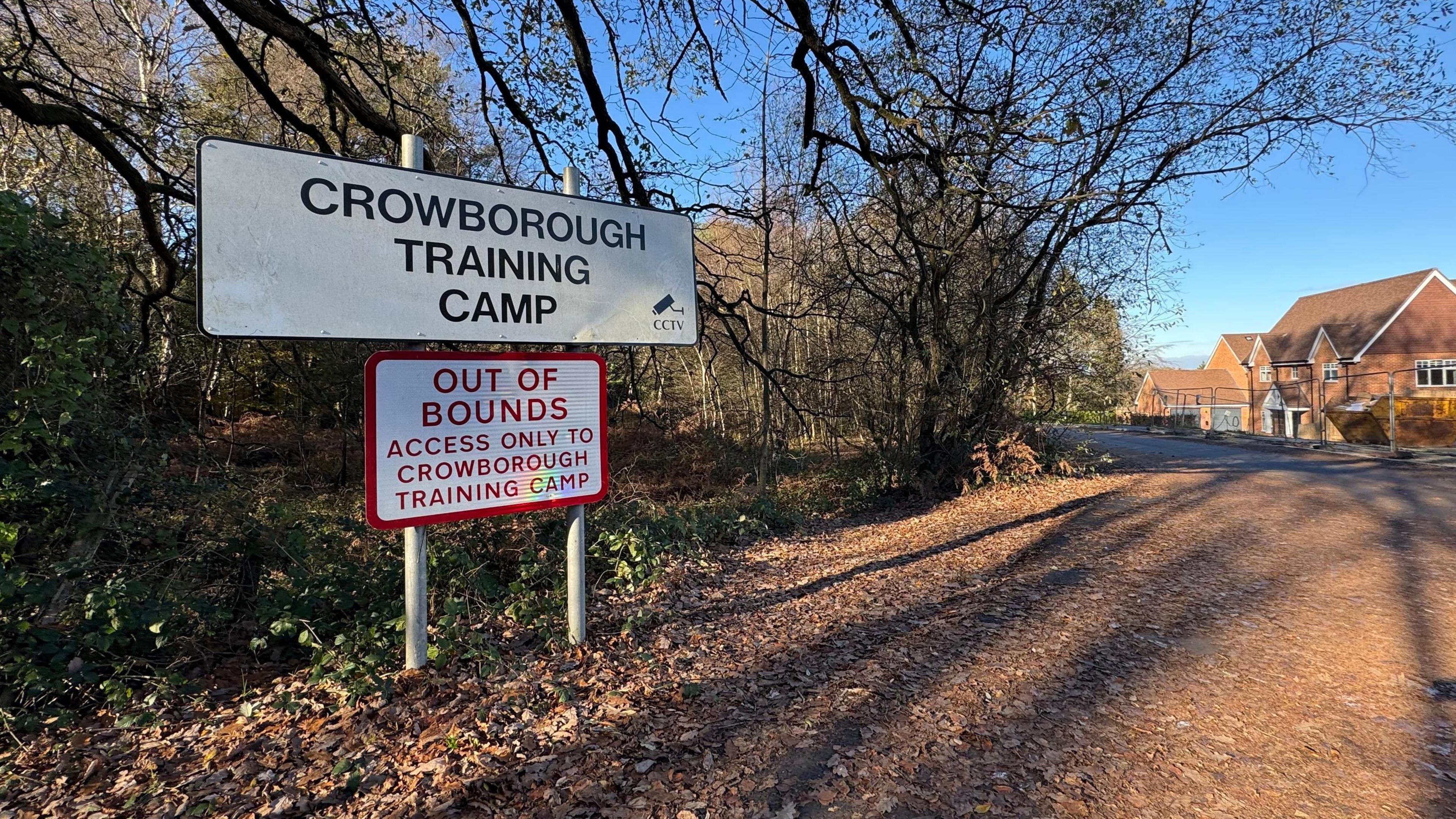 Under a blue sky and bare trees, a white sign marks the entrance to Crowborough Training Camp. Below the notice passers-by are warned the area is out of bounds. Next to the access road is a new housing estate. 