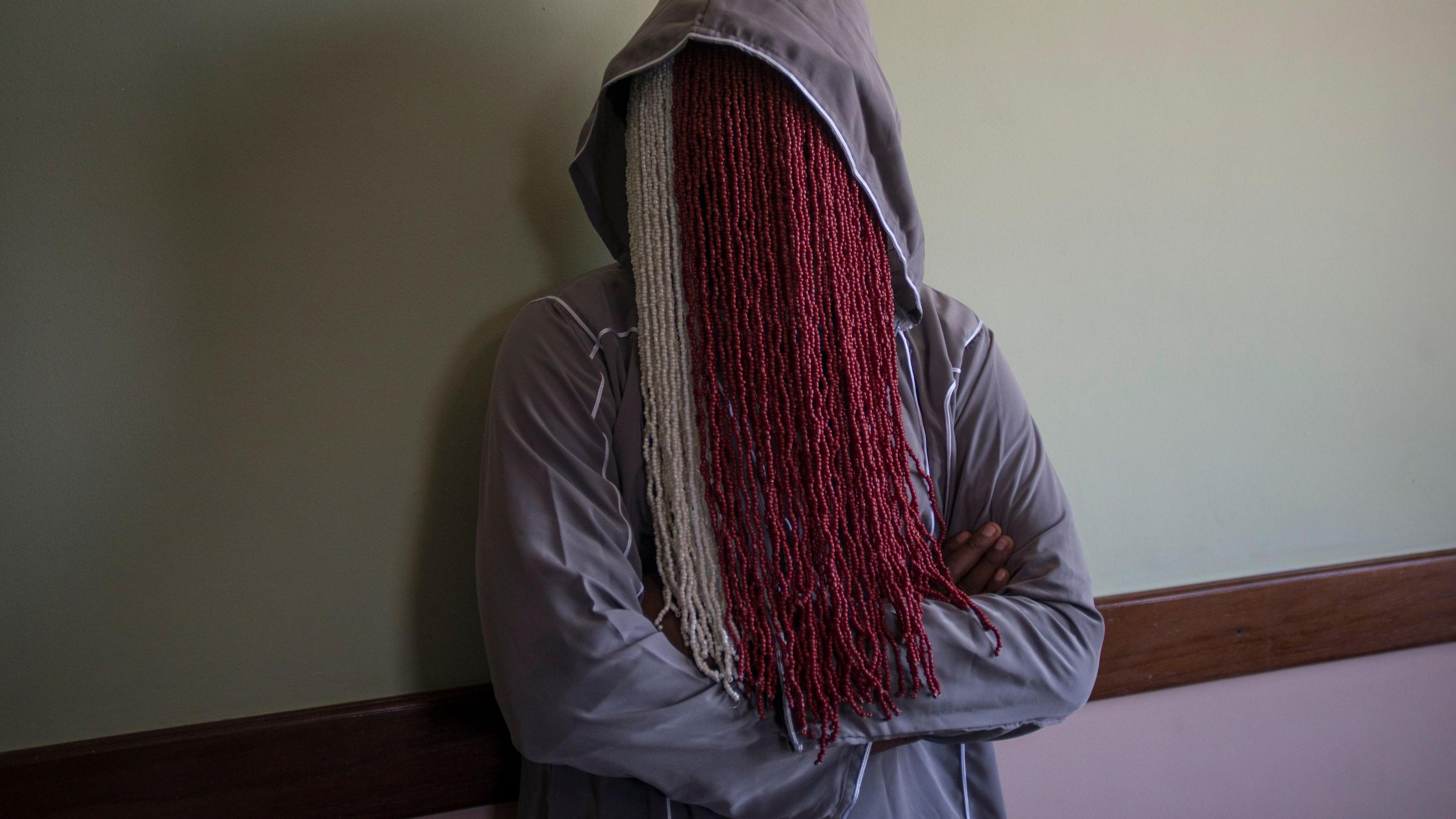 Anas Aremeyaw Anas is seen wearing a grey hoodie and with his arms crossed, while his face is totally obscured by a long white and red beaded mask which runs down to his chest