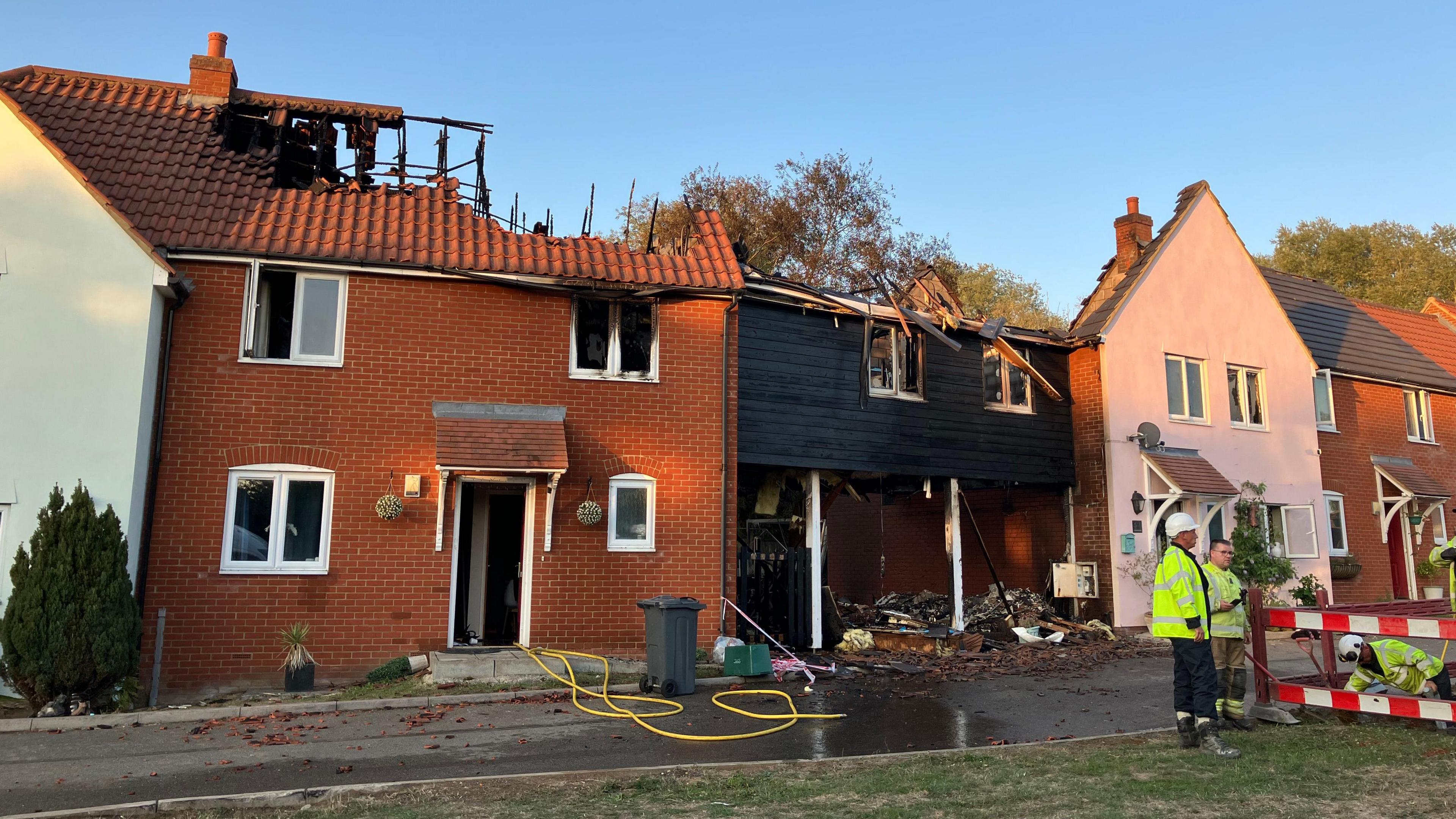 Three modern terraced houses on a new-build development have sustained fire damage, with their roofs totally destroyed. Men in high-vis yellow jackets are in the right of the picture. One is inside a hole in the ground, possibly working on a water main.