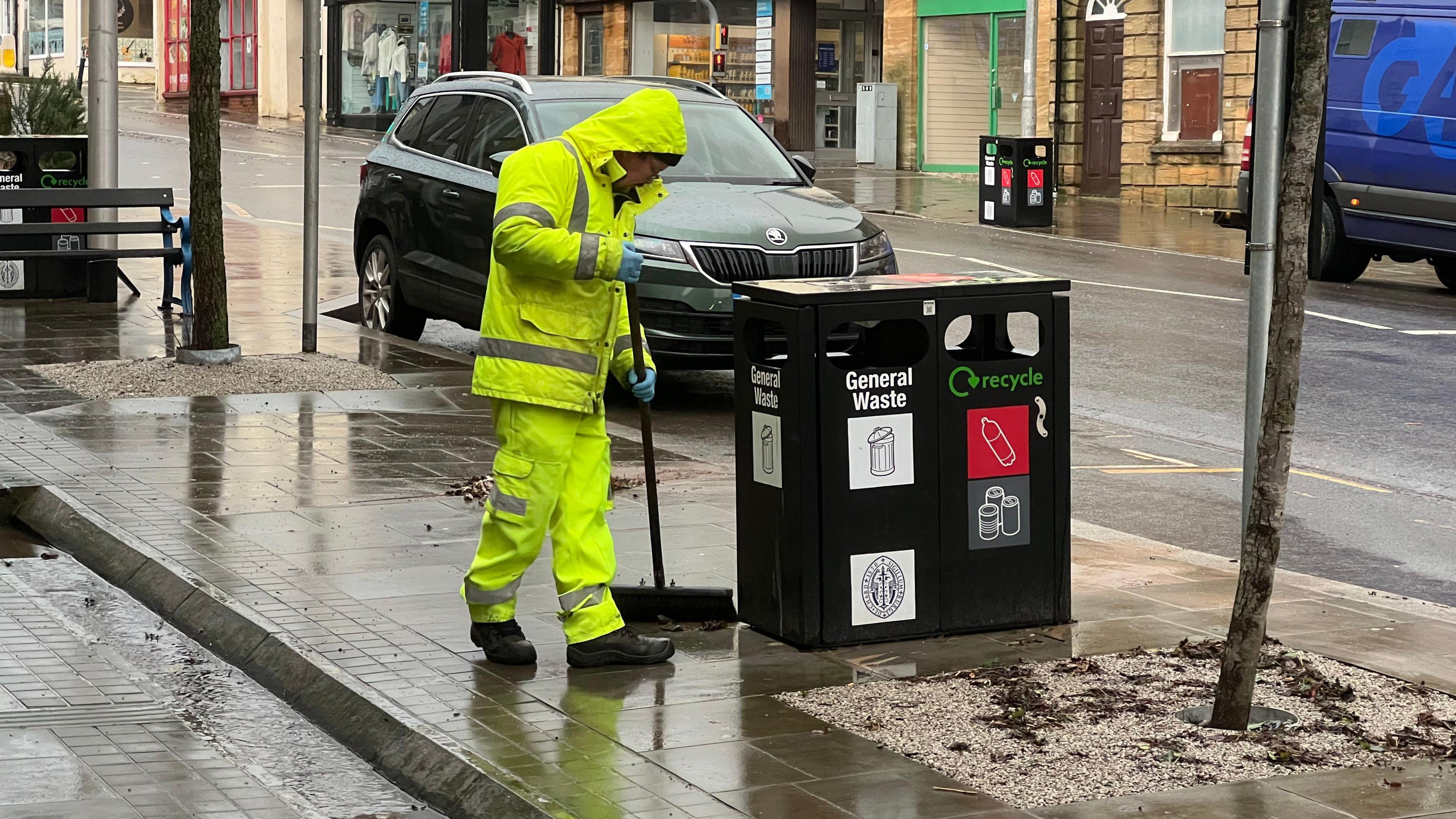 A street cleaner wearing a hi-vis jacket and trousers sweeping the pavement on a high street