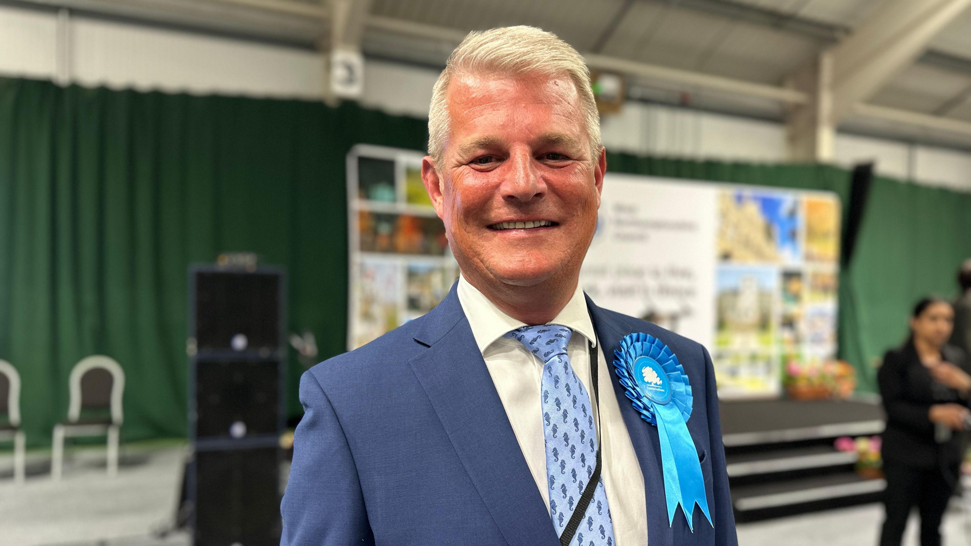 A head-and-shoulders shot of Stuart Andrew, a blond-haired man wearing a light blue suit, a beige shirt and a blue tie. He has a blue rosette pinned to his jacket. He is smiling at the camera. He is standing in a hall. The background is out of focus.