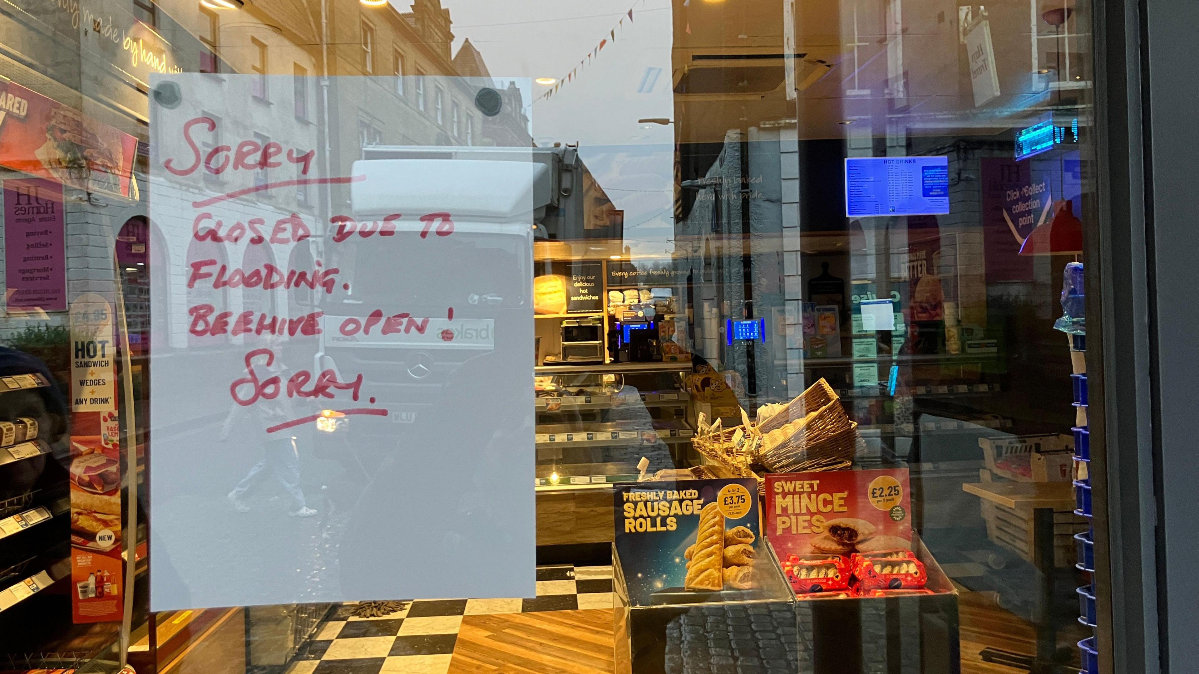 Window looking into a Greggs shop. Sign saying: "Sorry closed due to flooding. Beehive open. Sorry"