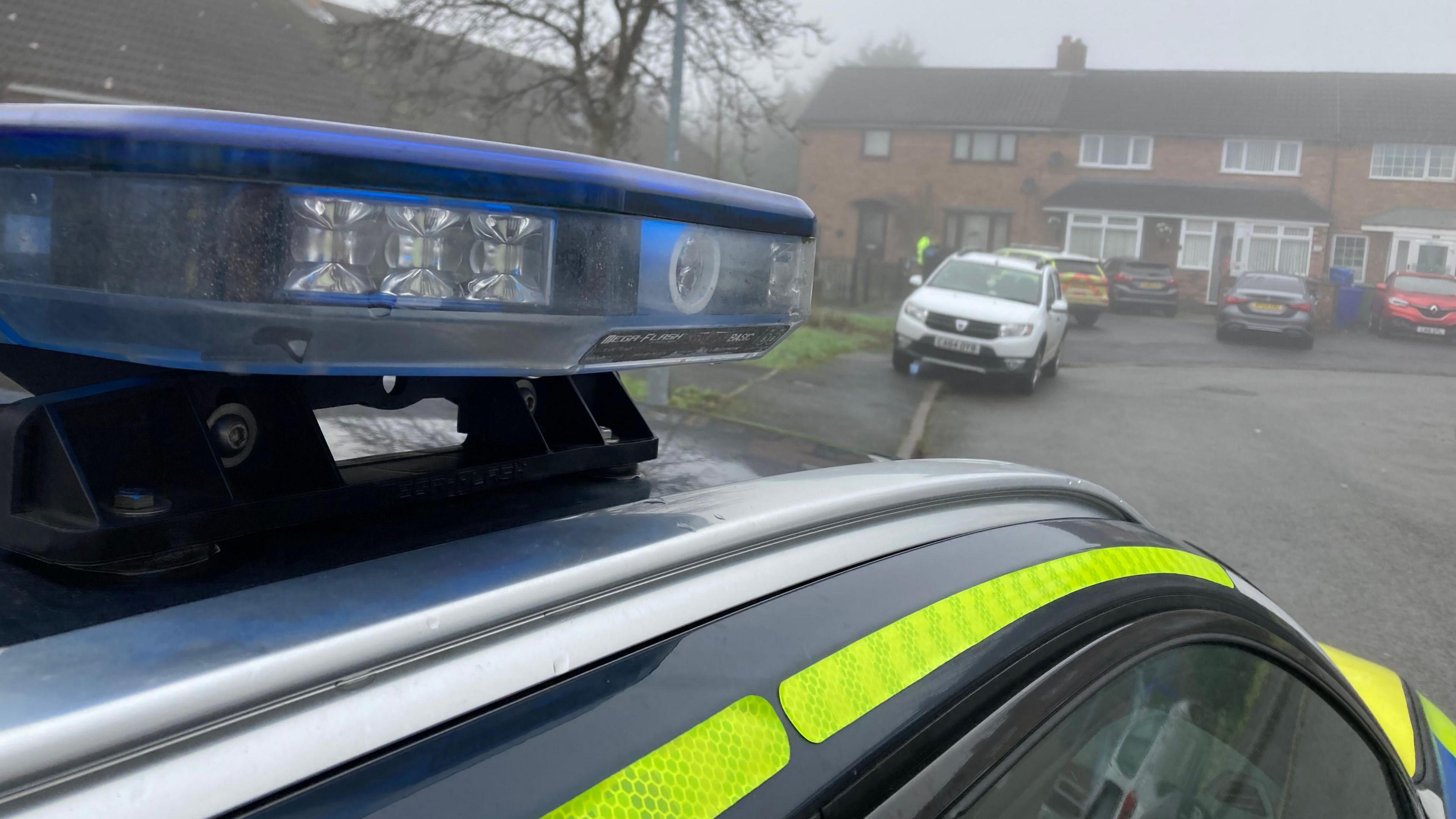 A Staffordshire Police car at the scene. Its siren can be seen on a foggy day and there is a police officer in the background near another car. 