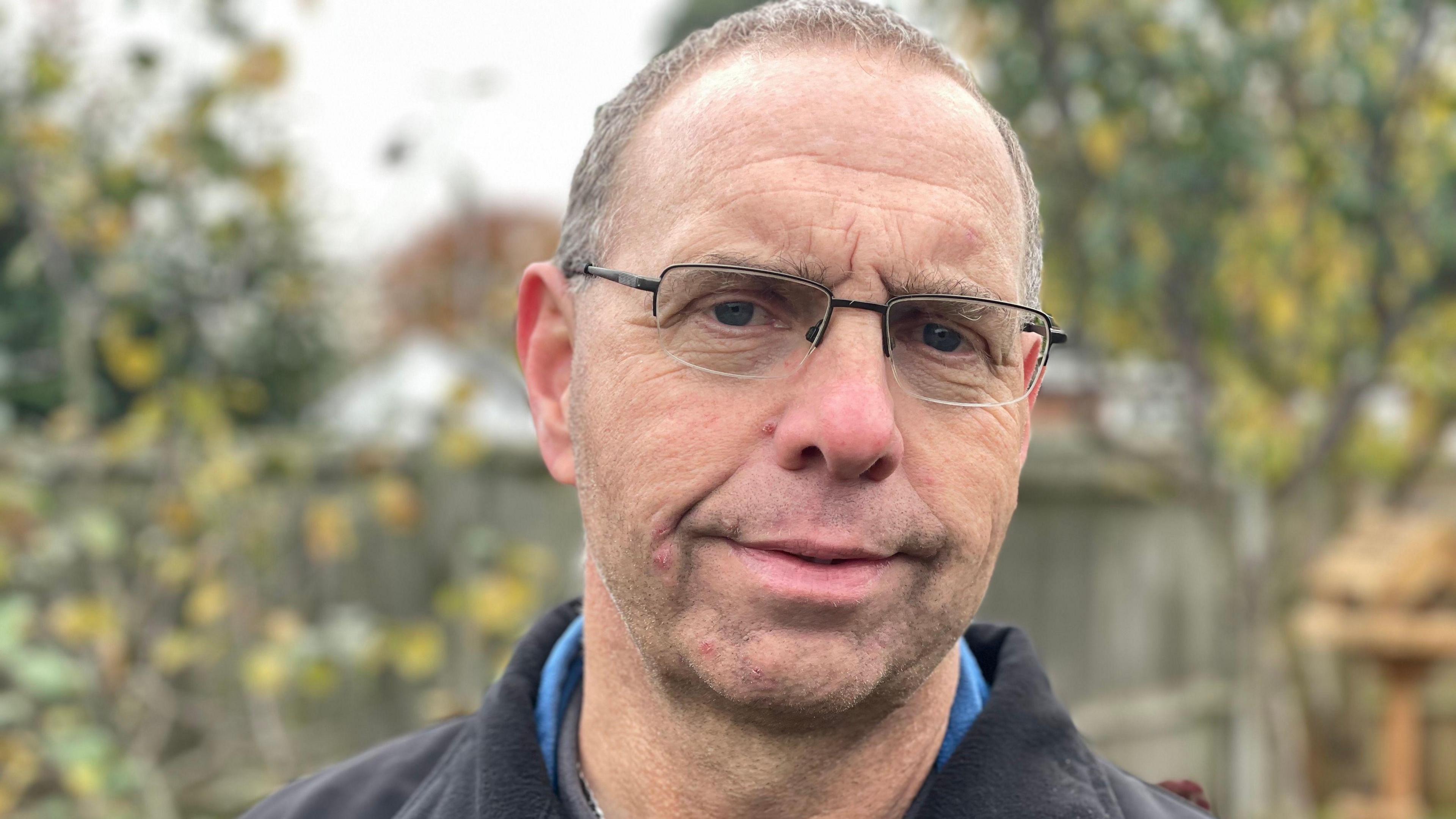 A man with glasses looks at the camera with blurred out trees and fencing in the background. He has short brown hair and is wearing a black jacket and blue t-shirt.