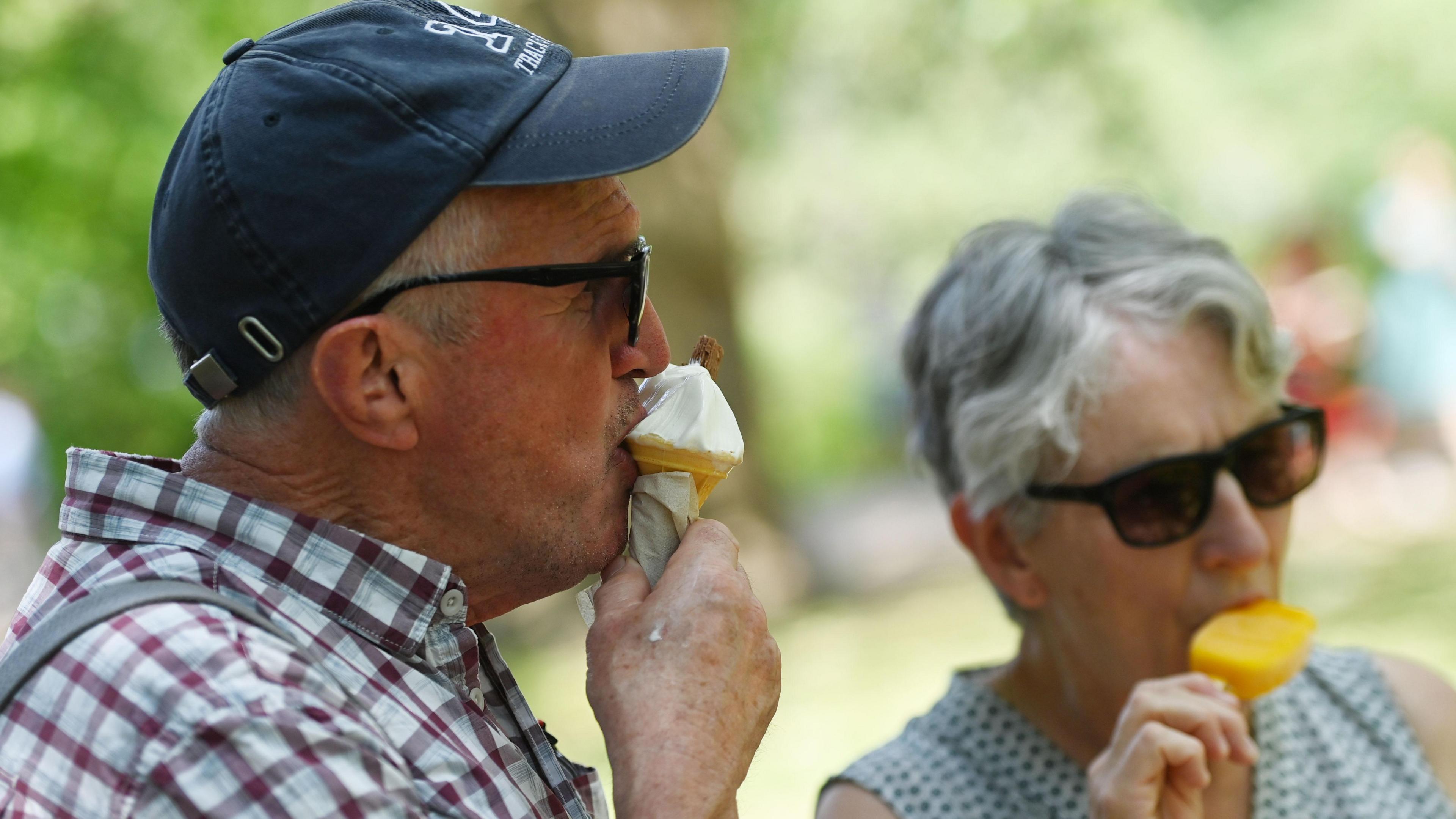 A man wearing a checked shirt and baseball cap tucks into an ice cream in a cone, with a chocolate bar on the top. A woman is enjoying an orange coloured iced treat in the background.