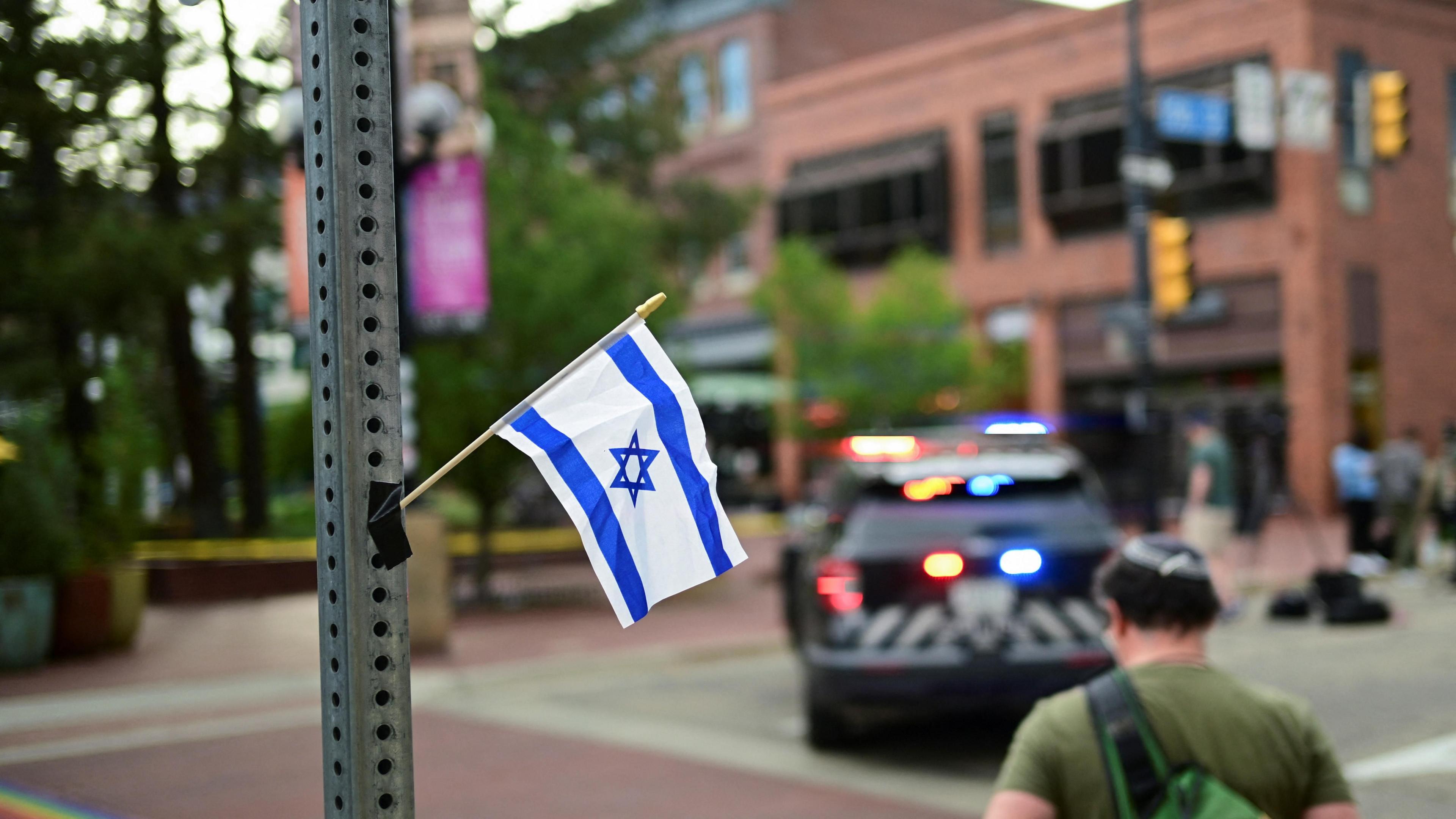 An Israeli flag is attached to a pole in the foreground as a police vehicle is shown in the background with red and blue lights flashing