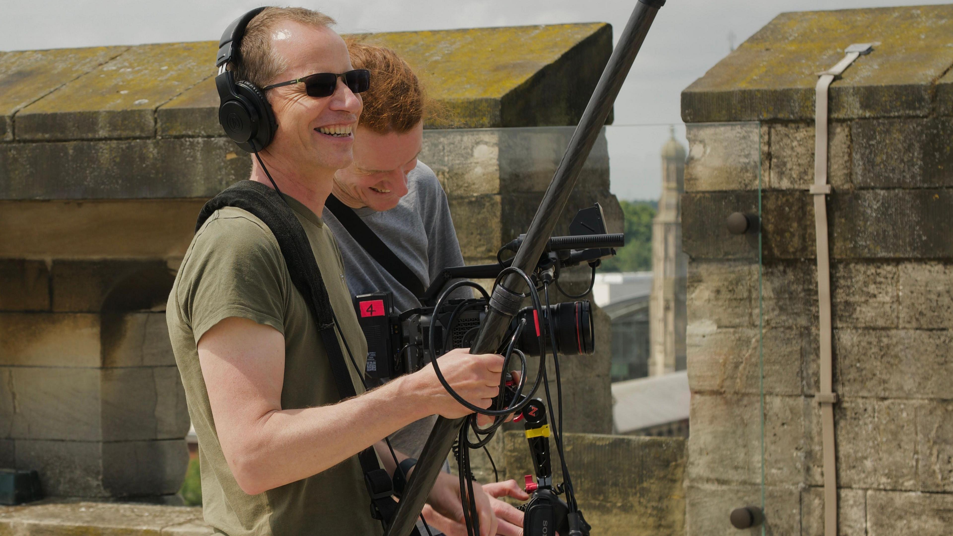 Two film crew members stand on the battlements. One is holding a boom sound mike and is smiling while wearing headphones and the other man is holding a video camera and looking down at it.