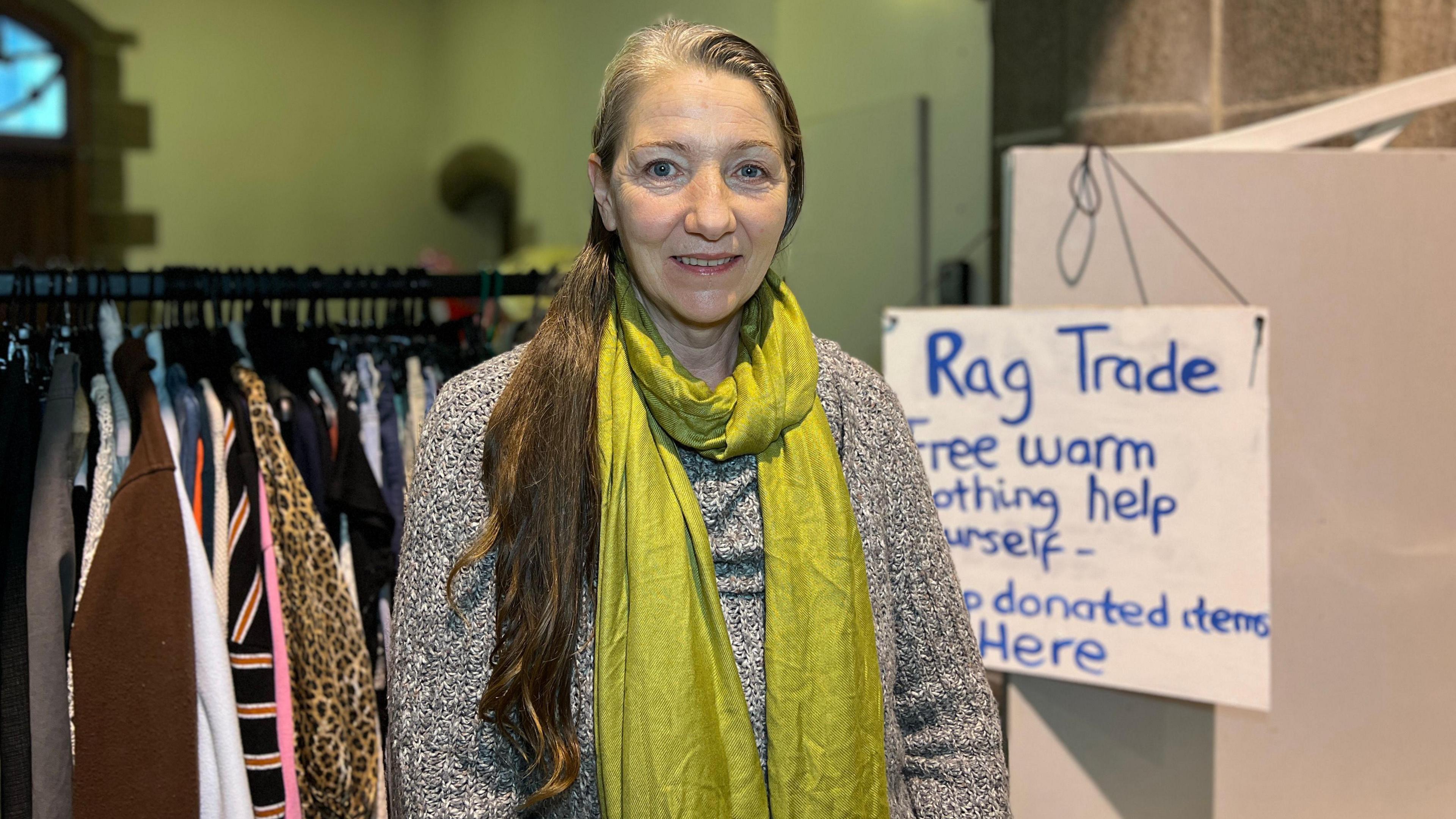 Kalina smiles at the camera as she stands in front of the clothes rail which has coats, jumpers and jackets on it. There is also a sign up for Rag Trade saying people can help themselves to the clothes and donate items as well. Kaline is wearing a grey and white jumper with a dull yellow scarf and she has long hair tied into a pony tail.