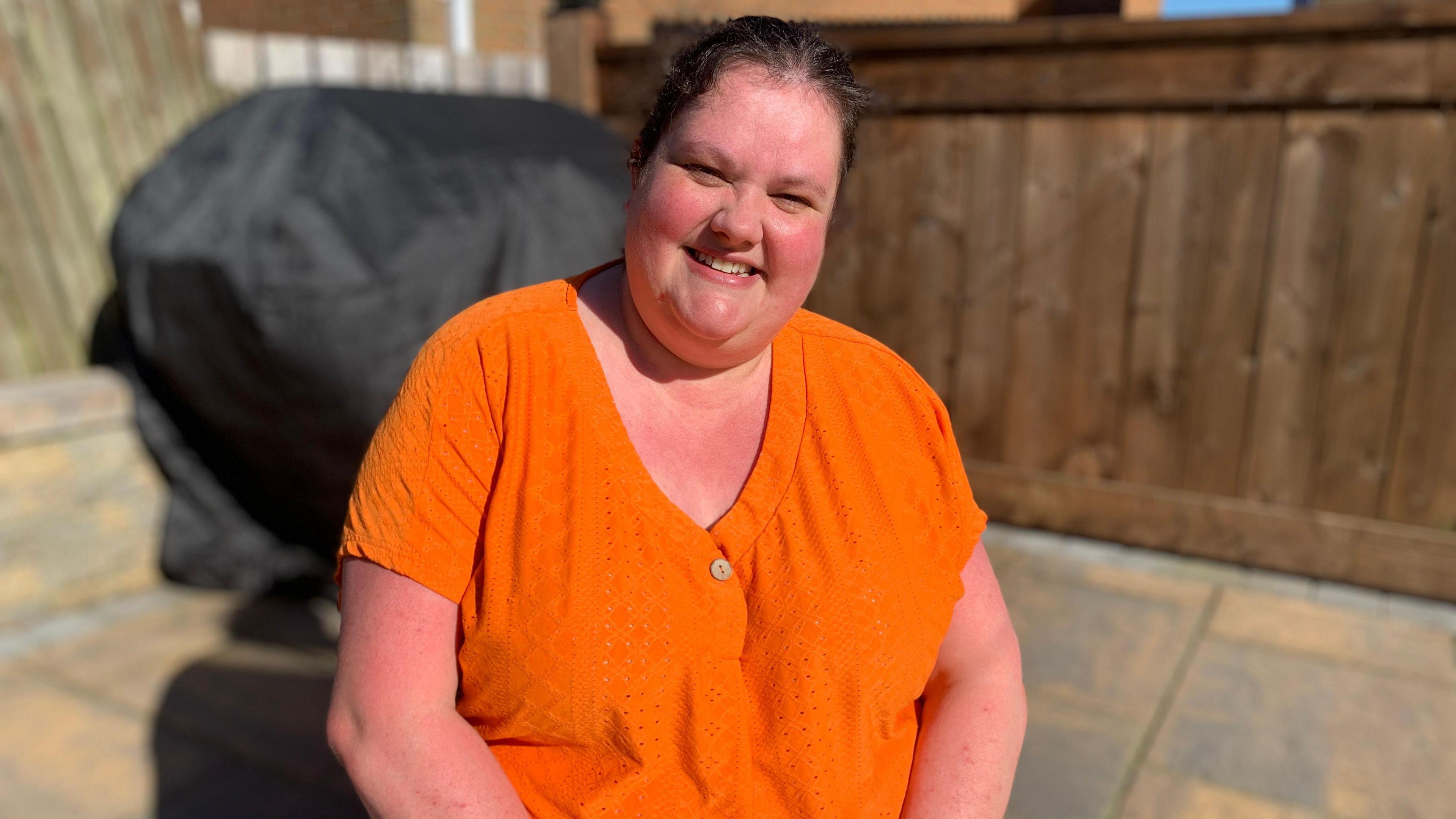 A woman in an orange top smiles at the camera. She has brown hair, tied back. It is a sunny day and she sits in front of a wooden fence.