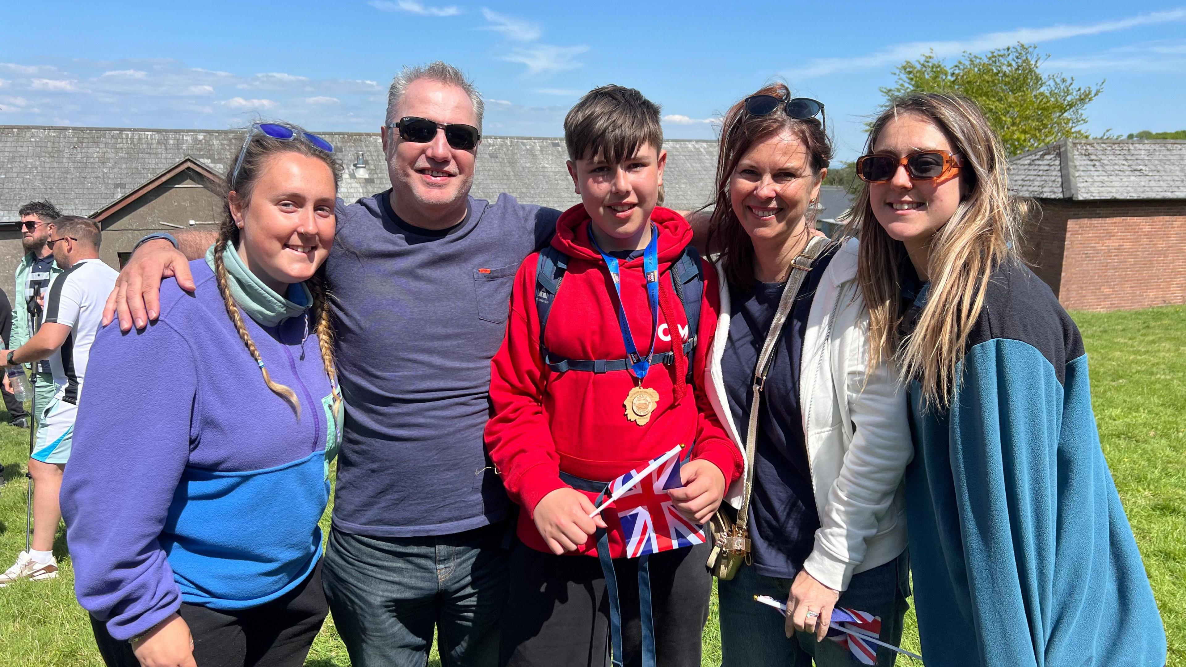 A family group of five people dressed in outdoor clothes. Ben is pictured in the middle wearing his medal over a red hoodie and holding a Union flag