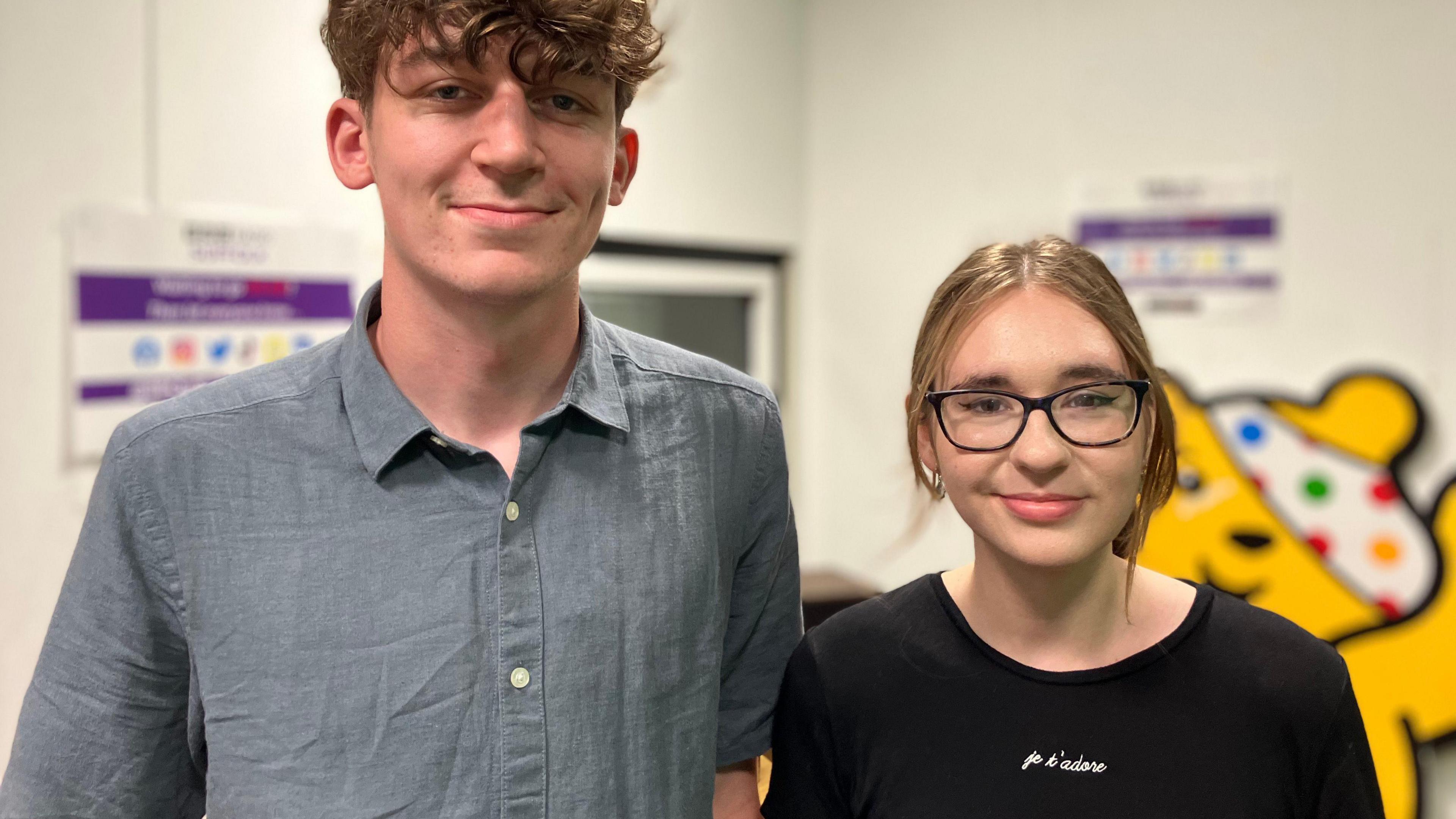A young man and a young woman standing in a room, smiling at the camera, with Pudsey Bear on the wall in the background.