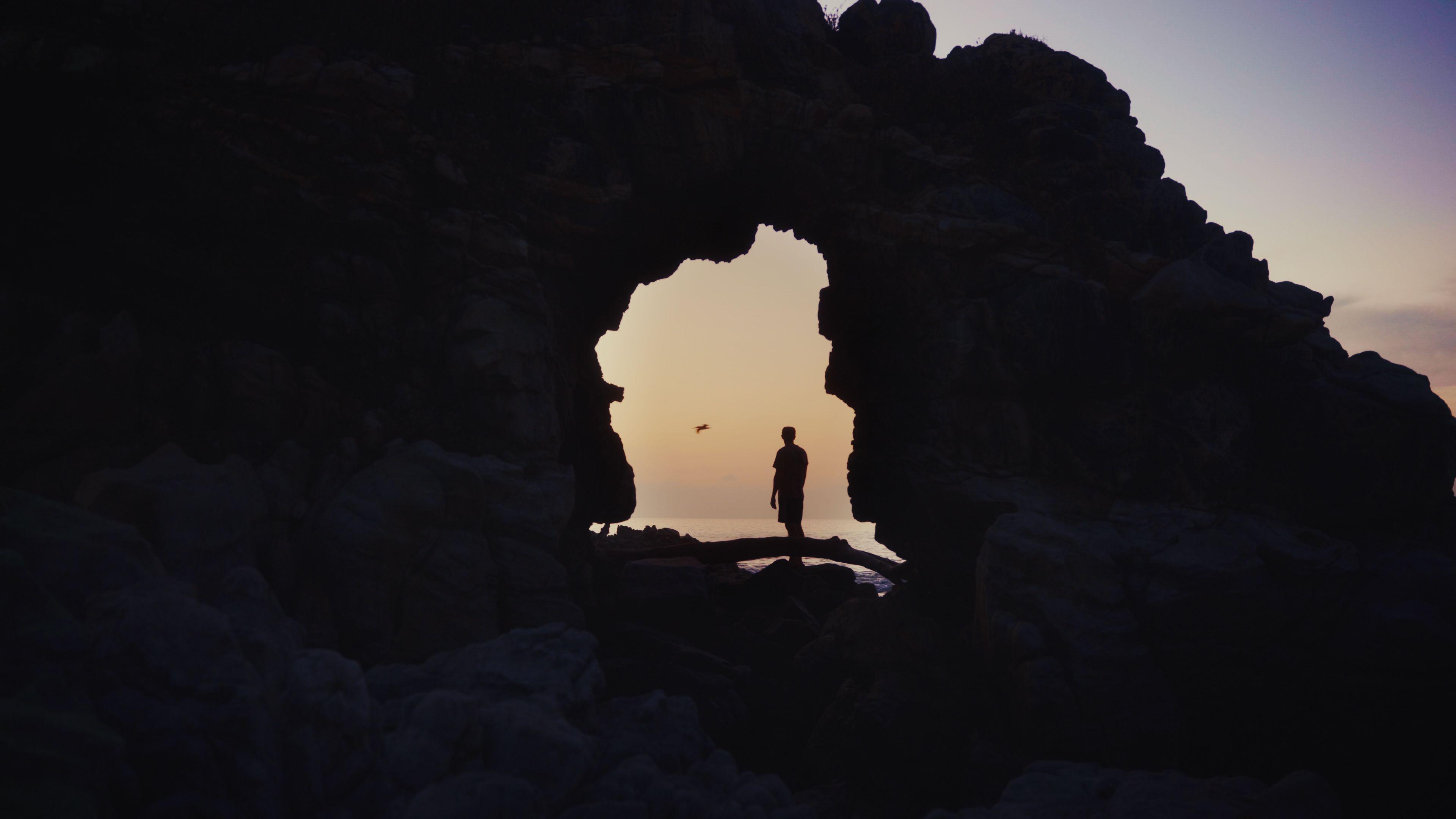 A man is stood in a natural hole near the sea. There is a bird in the distance and it's sunset.